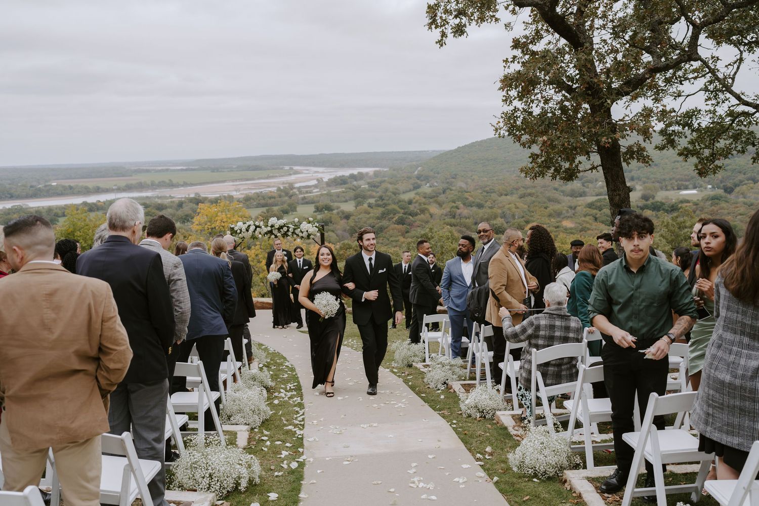 Wedding guests gather on a hillside path lined with white chairs overlooking a scenic valley.