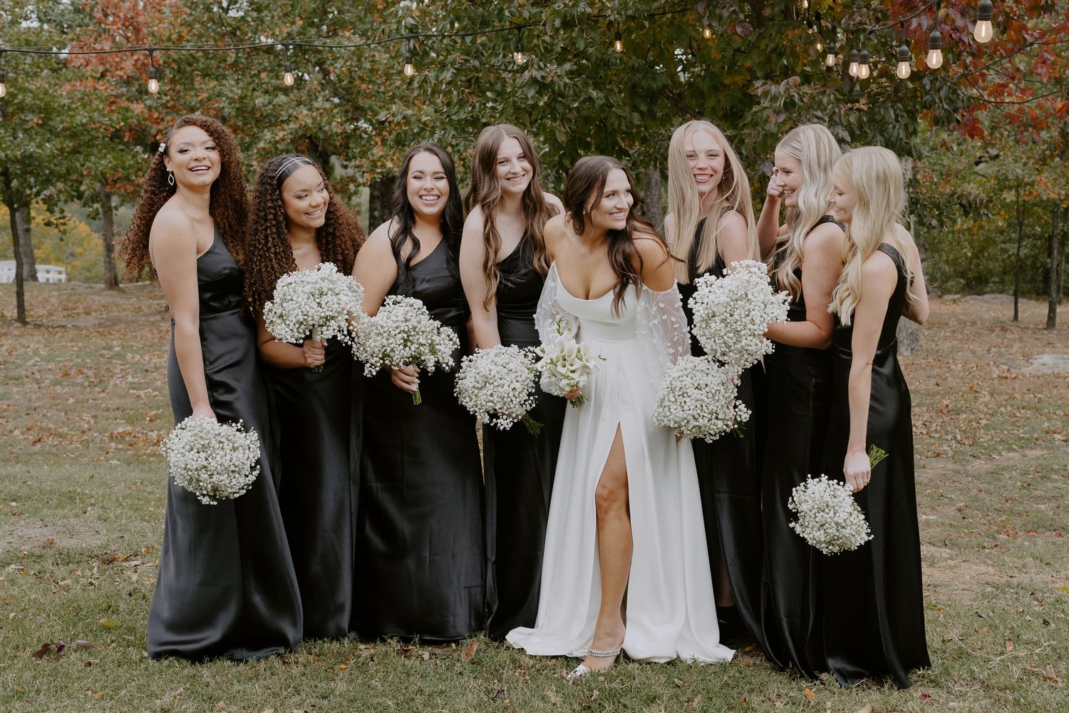 Wedding party members in black attire gather for photos against a backdrop of fall trees.
