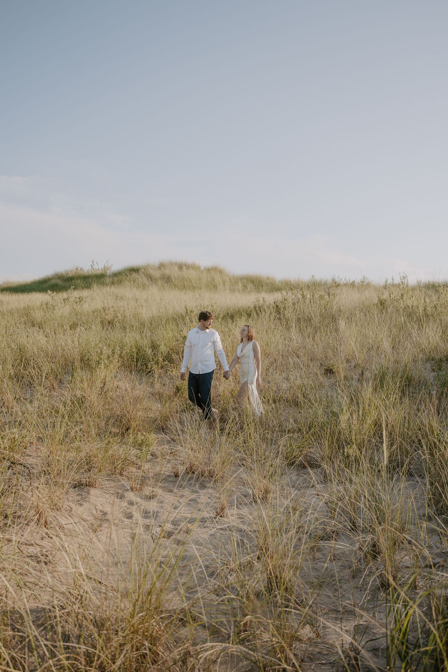 Two people walk hand in hand through coastal dunes on a hazy afternoon.