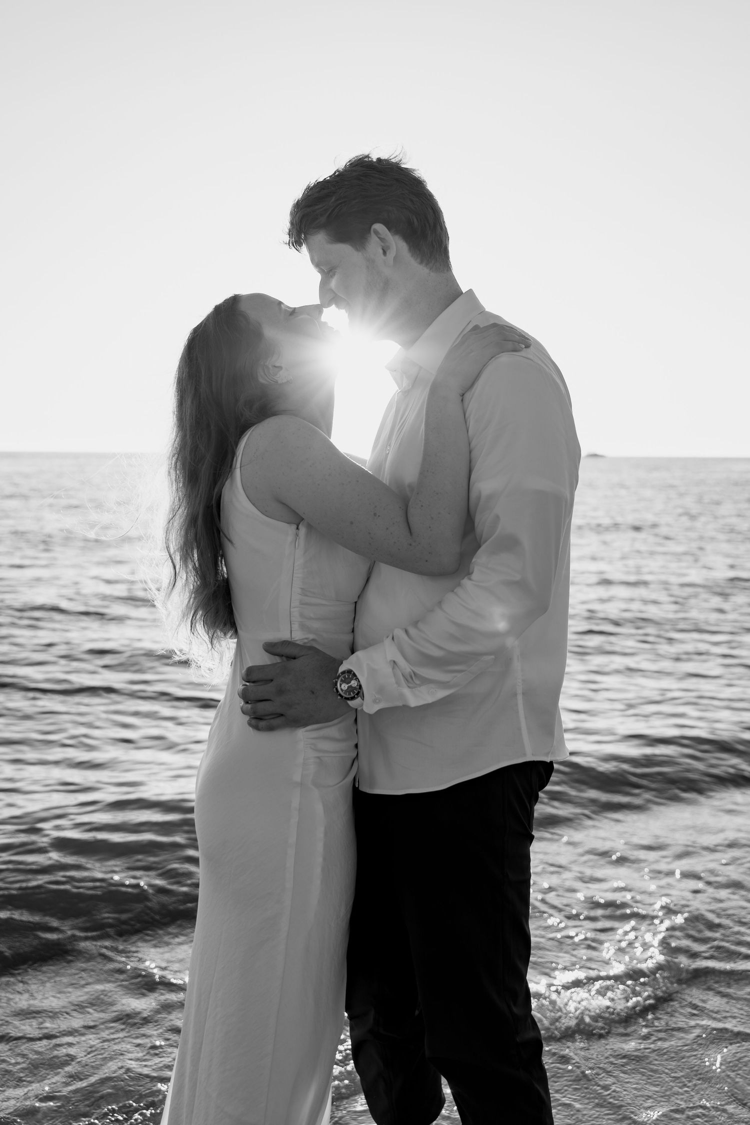 Backlit couple in formal wear sharing tender moment on beach at dusk.