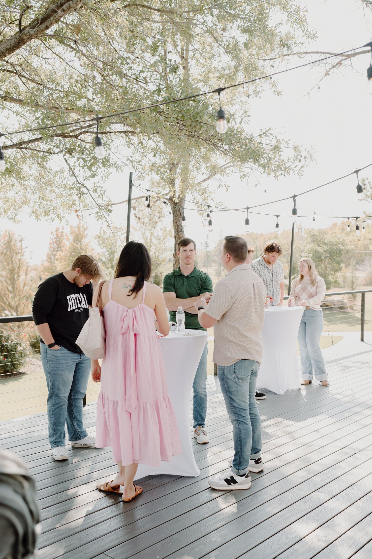 People mingling on a covered wooden deck with string lights and pink accents.