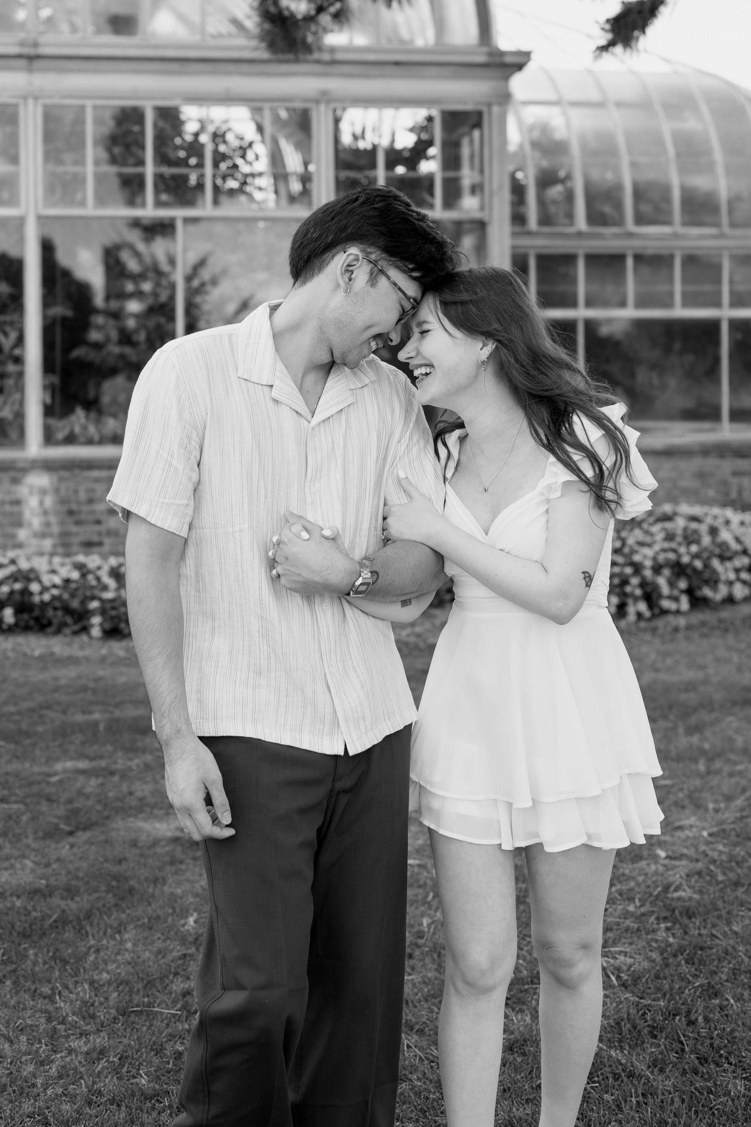 A couple shares affectionate moments during their photo session by a greenhouse with blooming flowers.