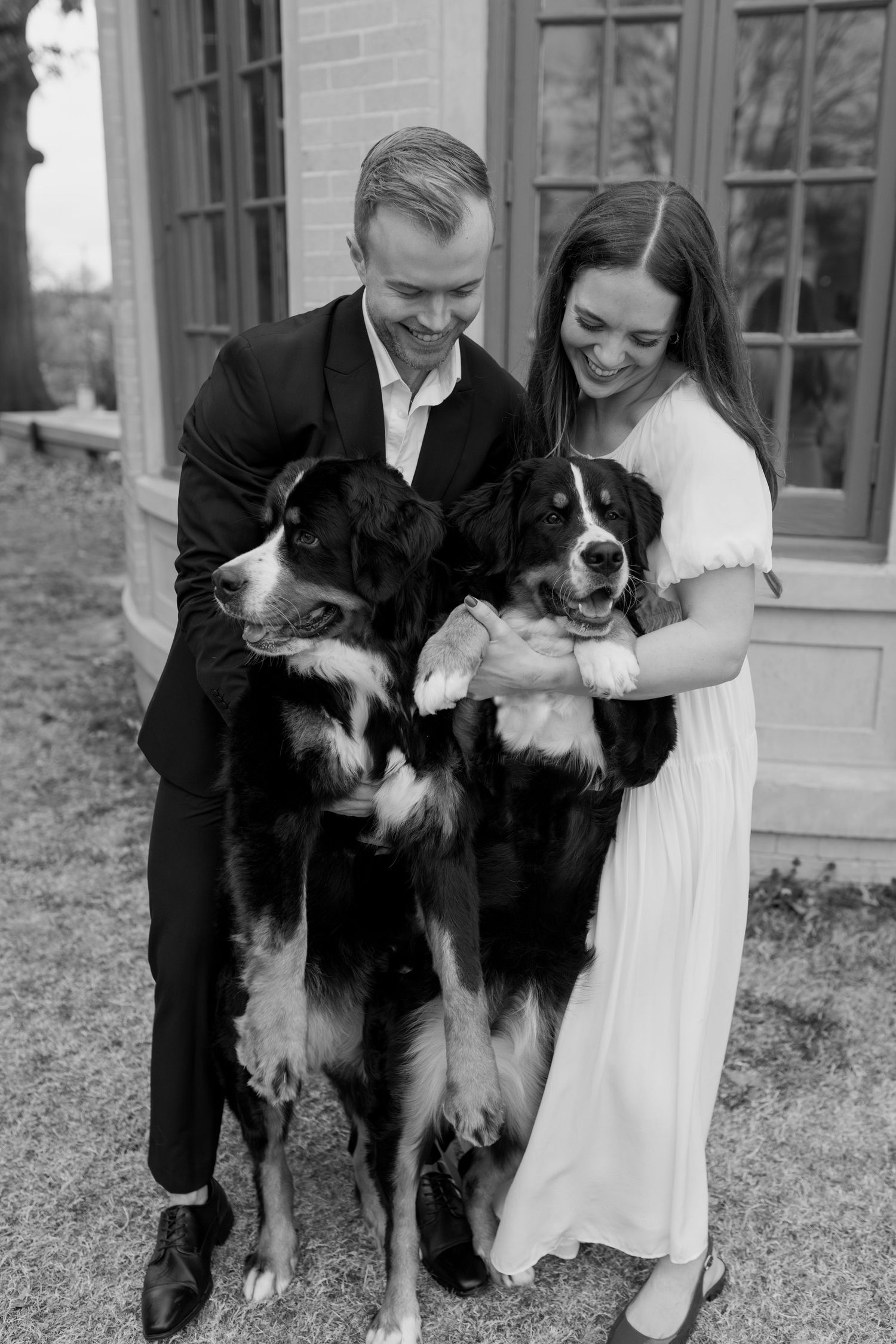 A couple poses with their two black and white Bernese Mountain Dogs outside a brick building.