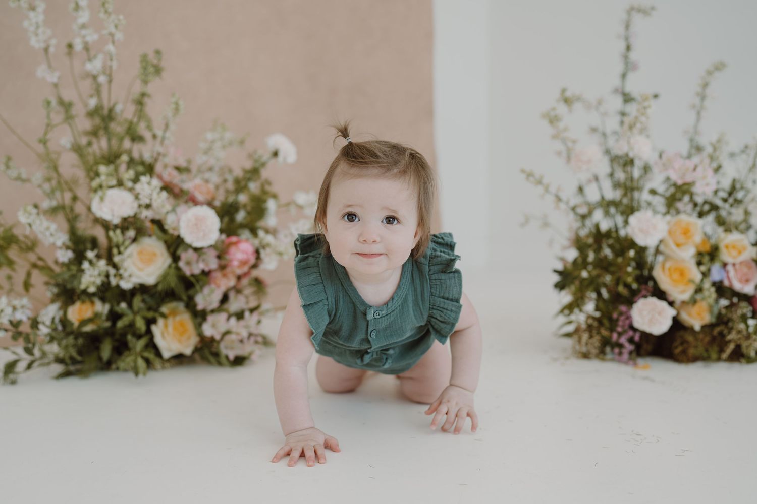 A small child in a green ruffled outfit crawls on a white floor with floral arrangements in the background.