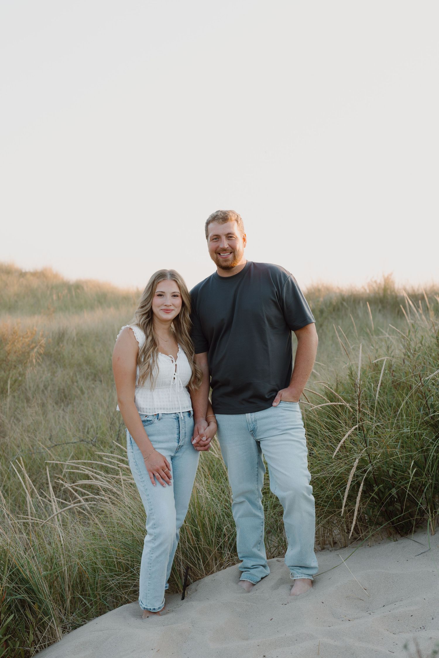 A couple in matching light denim and dark tops poses together on sandy dunes at sunset.