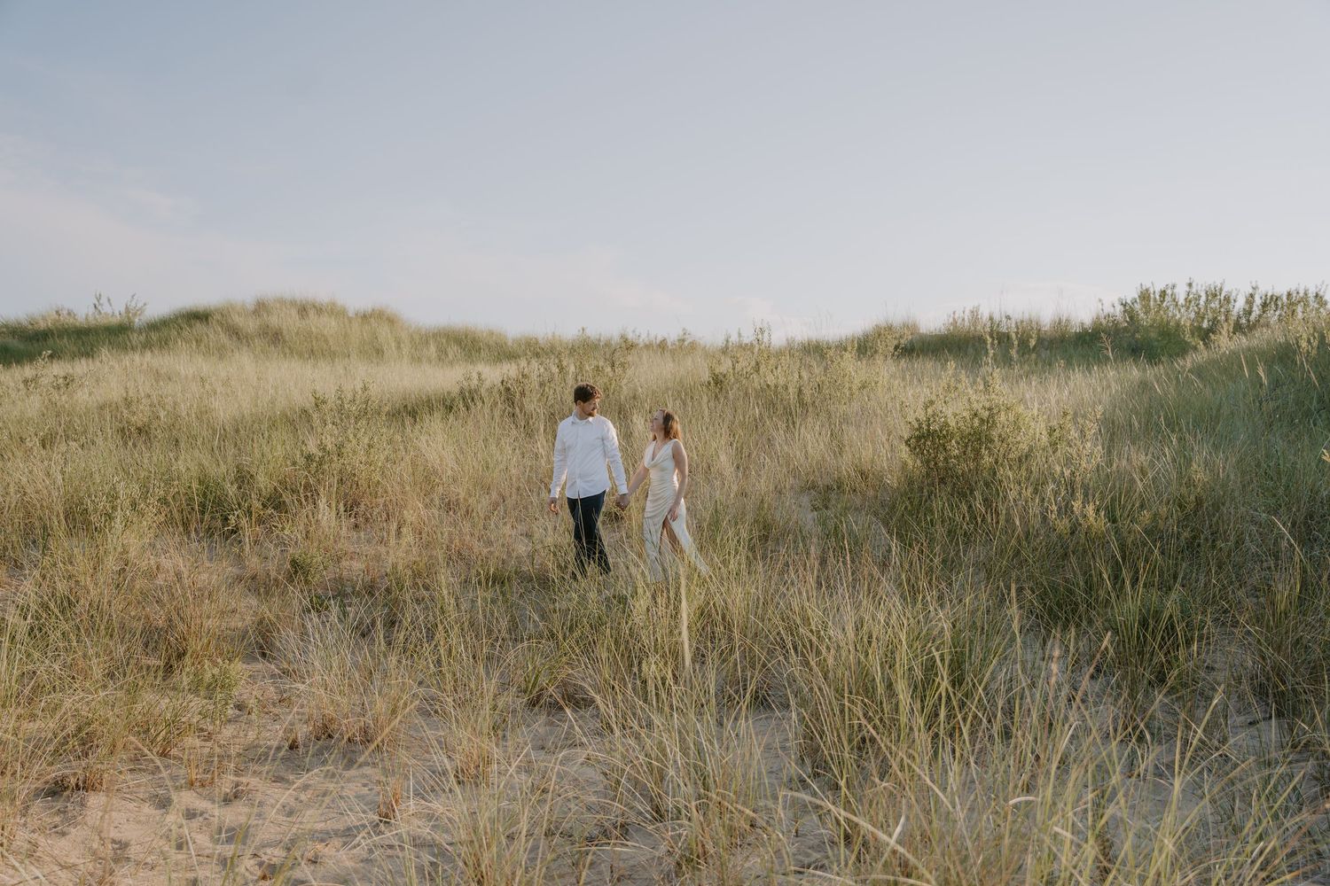Two figures walk together through coastal dune grass at sunset.