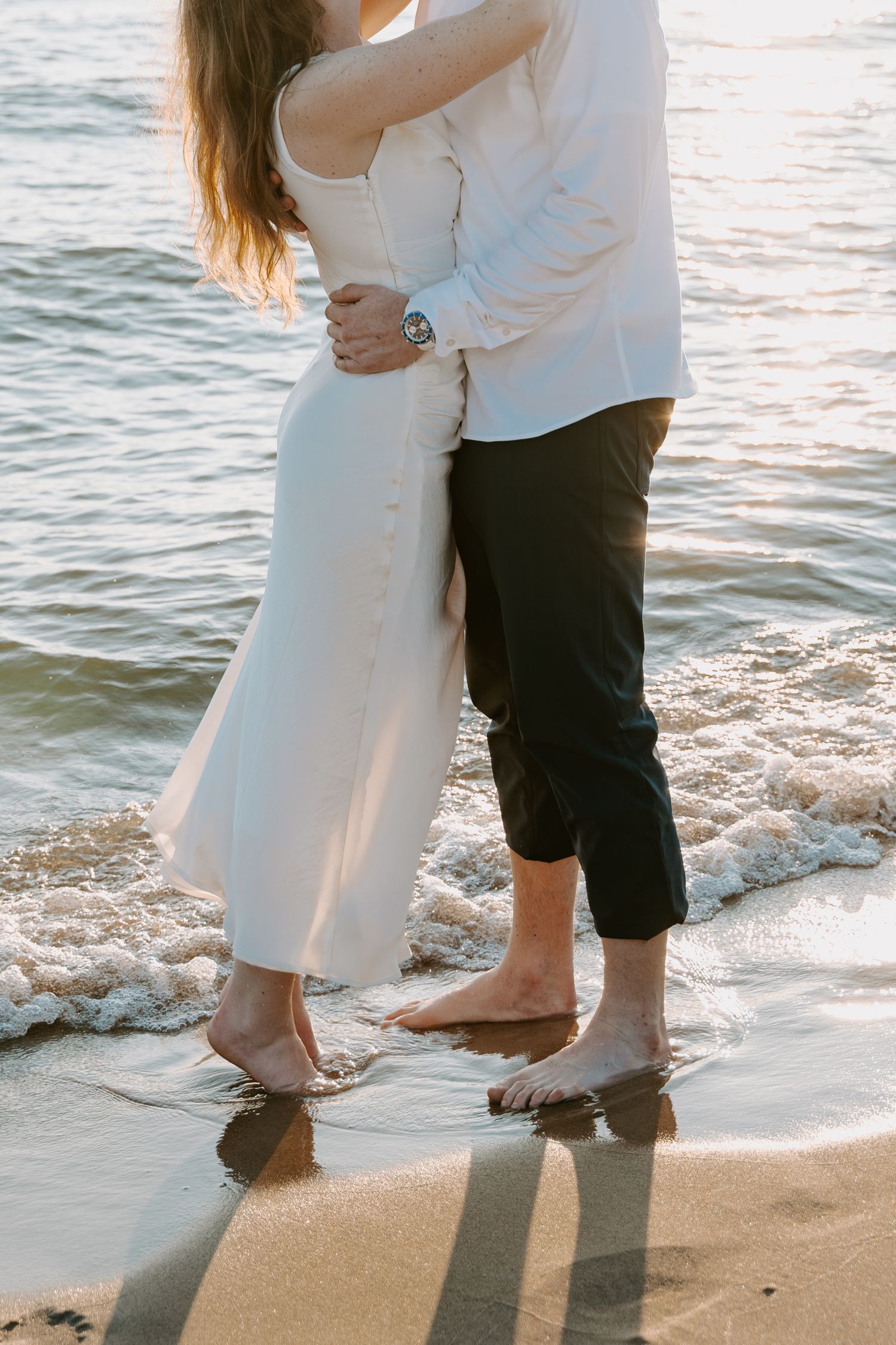 Close-up of a couple's feet touching in the sand as waves lap the shore.