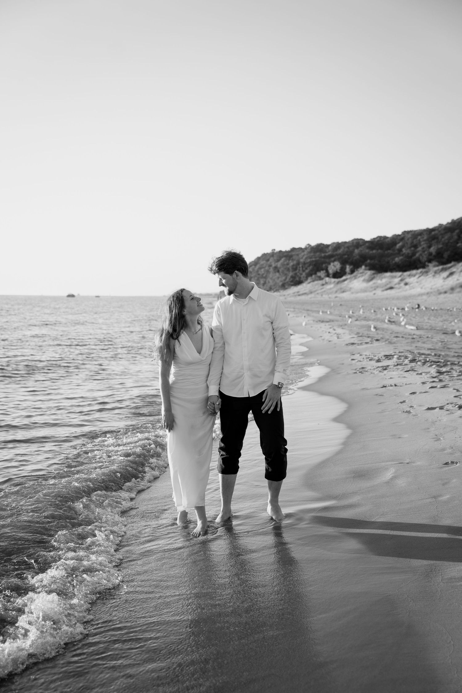 Black and white photo of couple walking along shoreline with waves lapping at their feet.