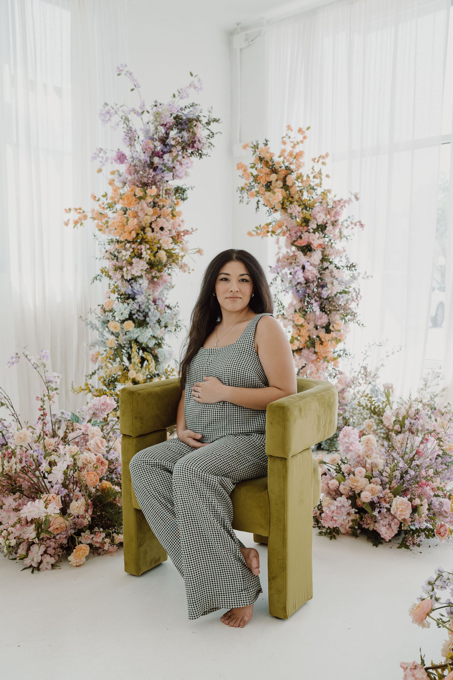 Maternity portrait featuring woman in striped jumpsuit with floral arrangements and white backdrop.