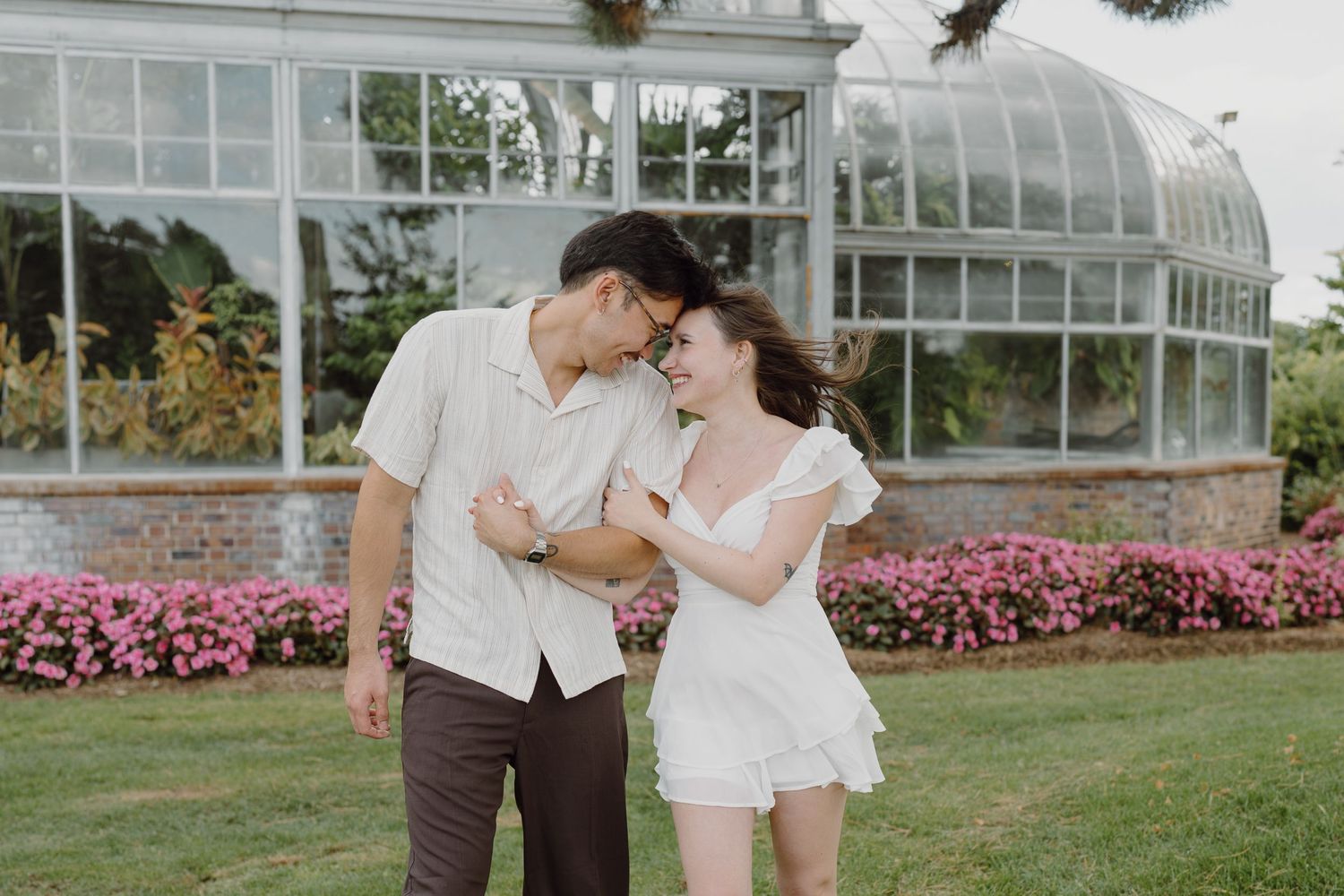 A couple shares an intimate moment in front of a greenhouse with pink flowers blooming nearby.