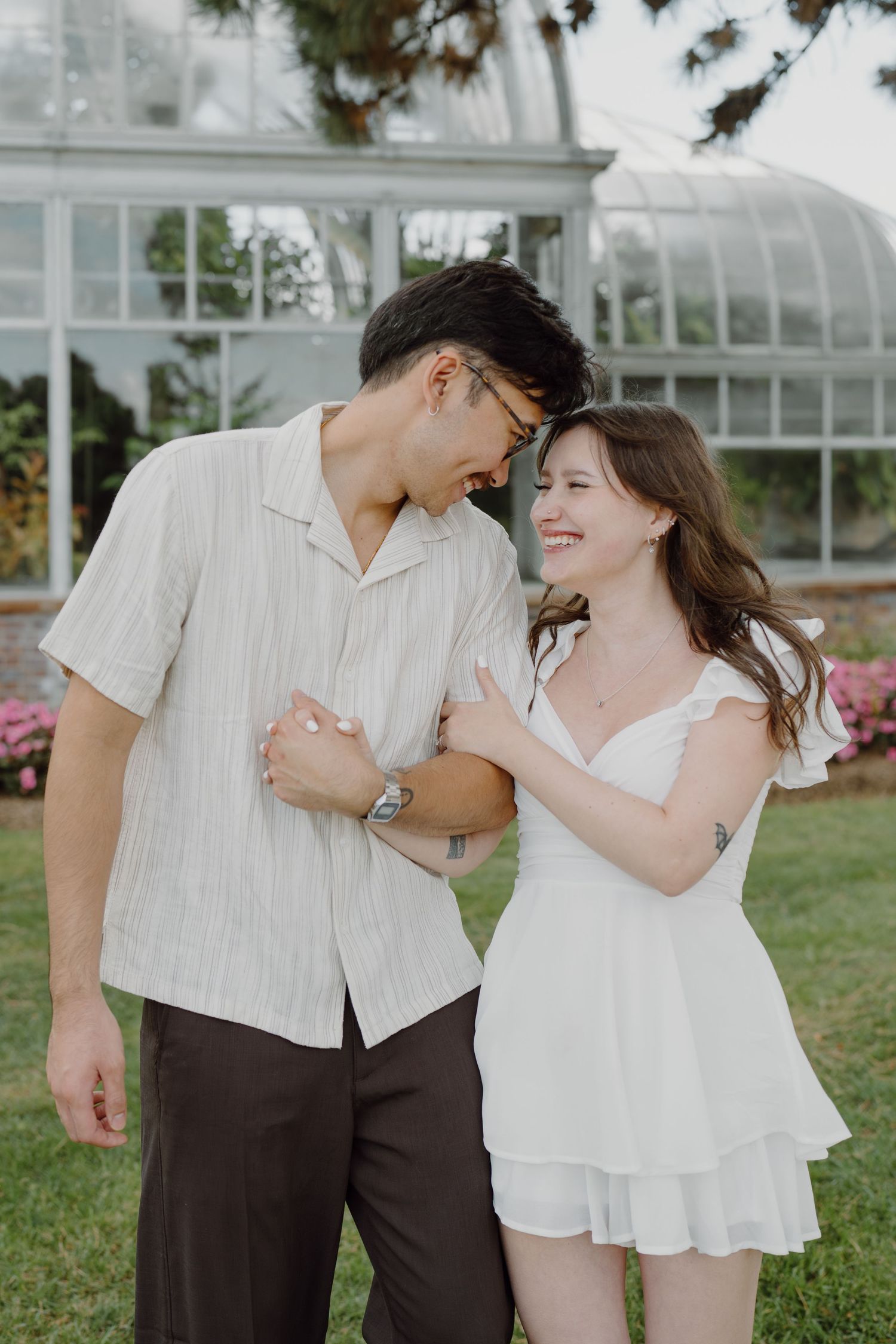 A couple dances and embraces near a greenhouse with pink flowers in the background.