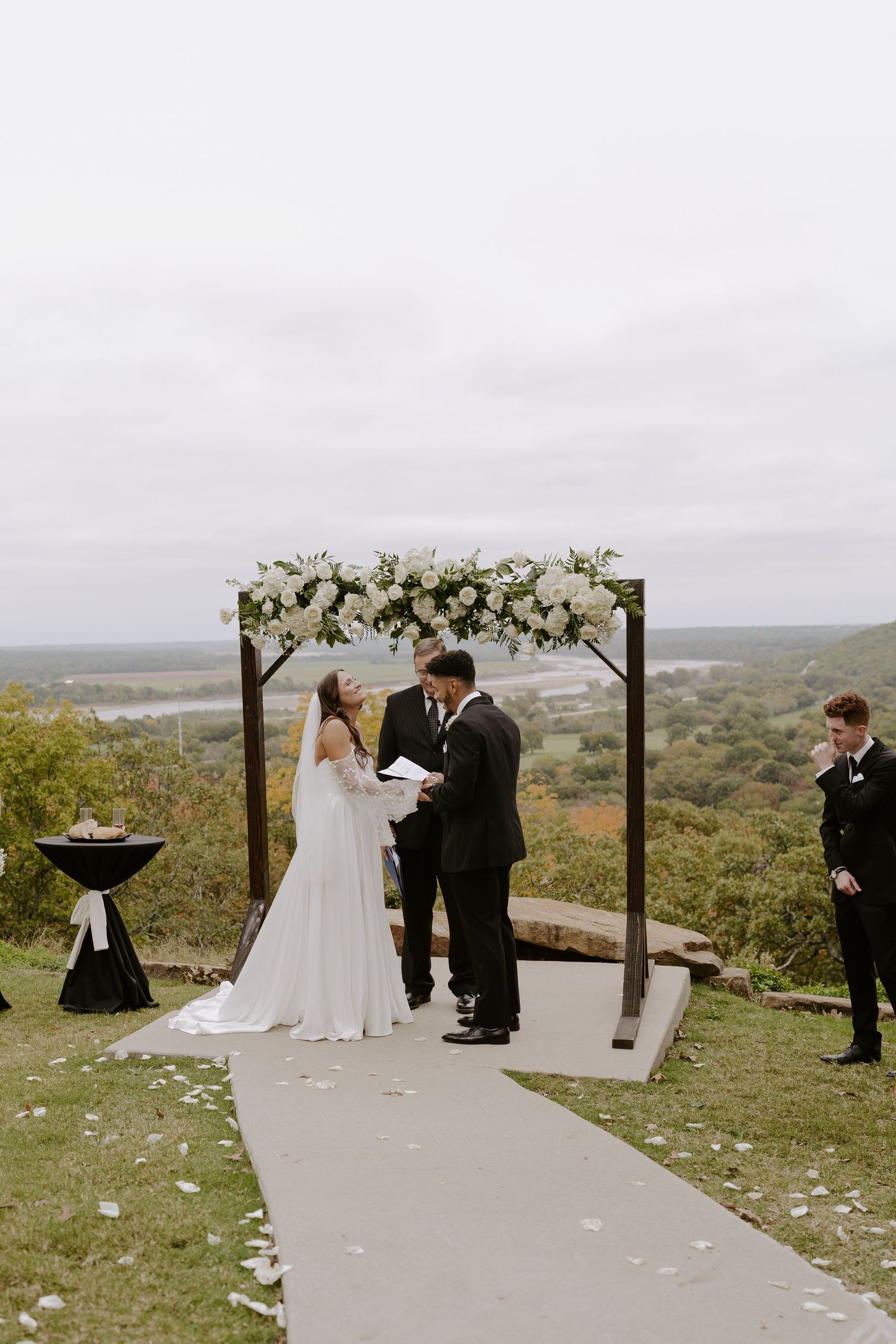 Wedding ceremony taking place on elevated platform with white floral arch and scenic valley backdrop.