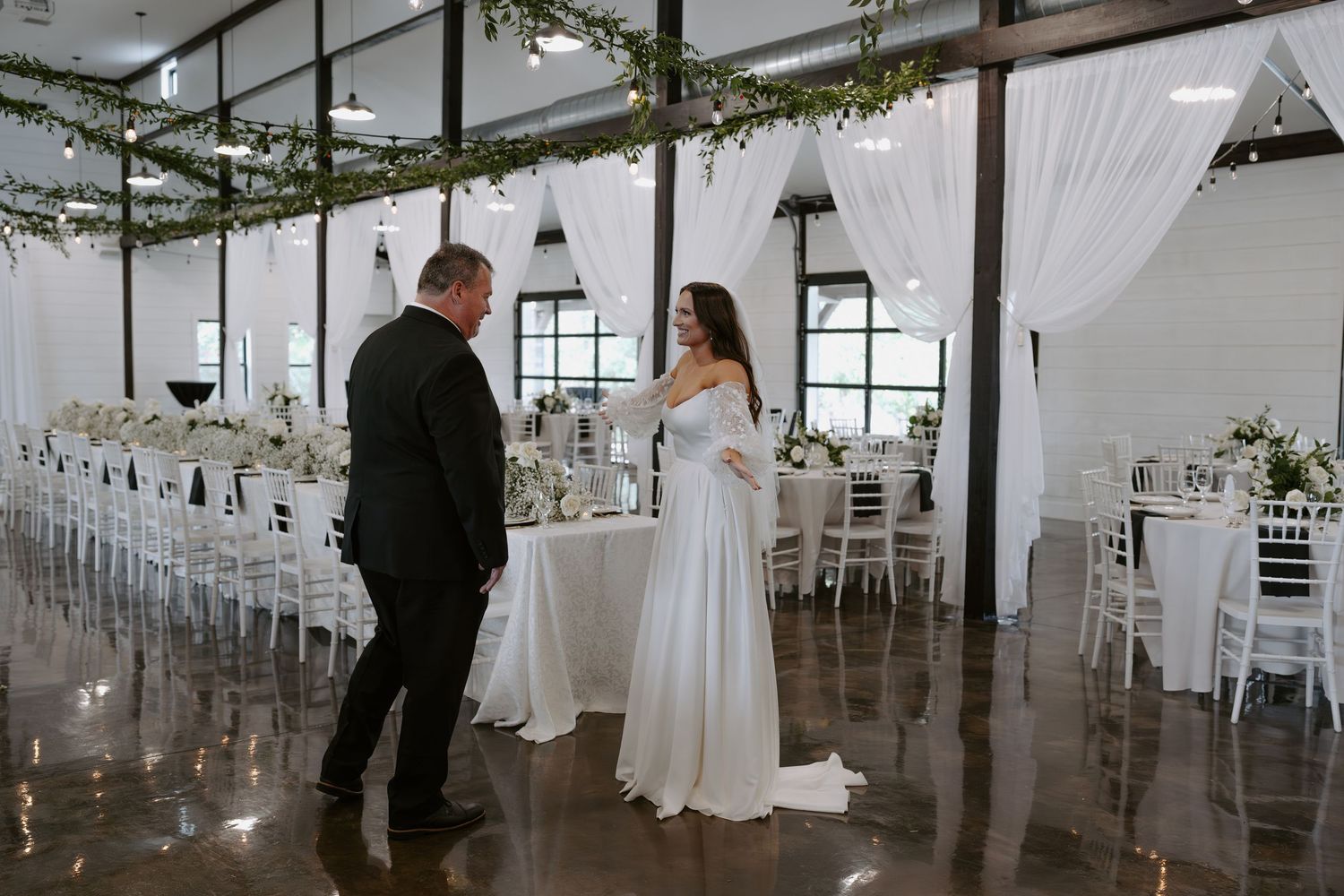 A couple stands face to face in an elegant wedding reception hall with white decor and greenery.