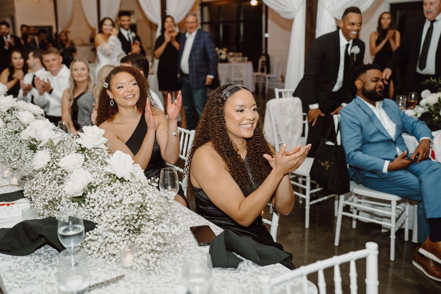 Wedding guests seated at tables with white floral centerpieces reacting during reception.