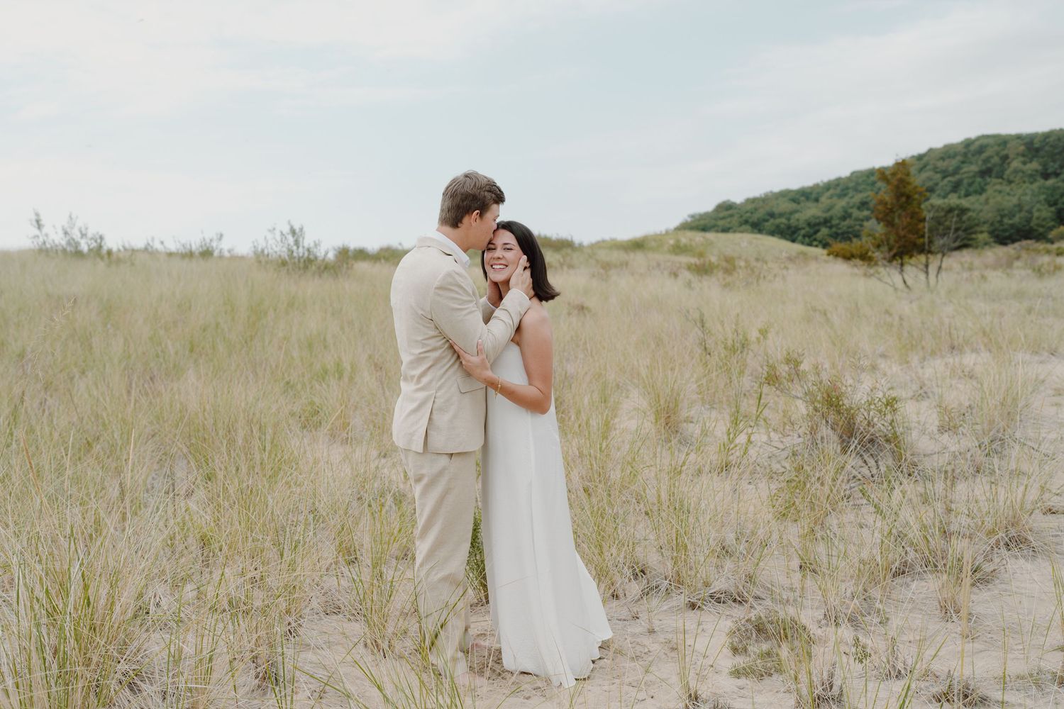A romantic beach wedding photo shoot with a couple embracing on sandy dunes surrounded by beach grass.