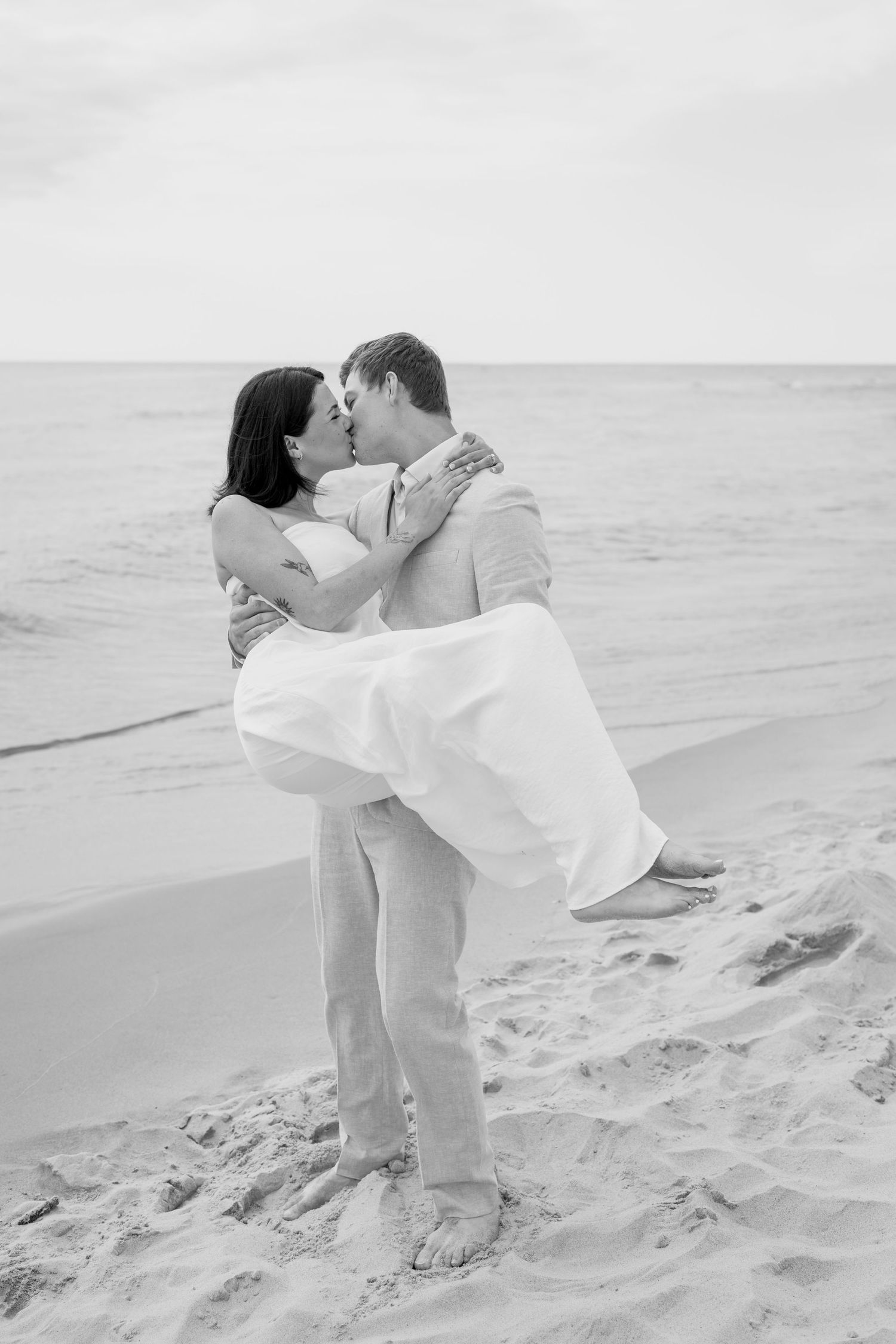 Black and white photo of couple embracing on sandy beach dunes during romantic moment.