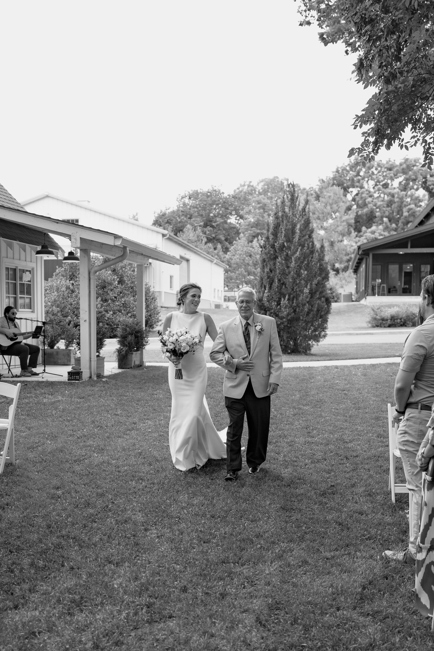 A black and white photo captures a wedding processional on a manicured lawn with buildings in background.