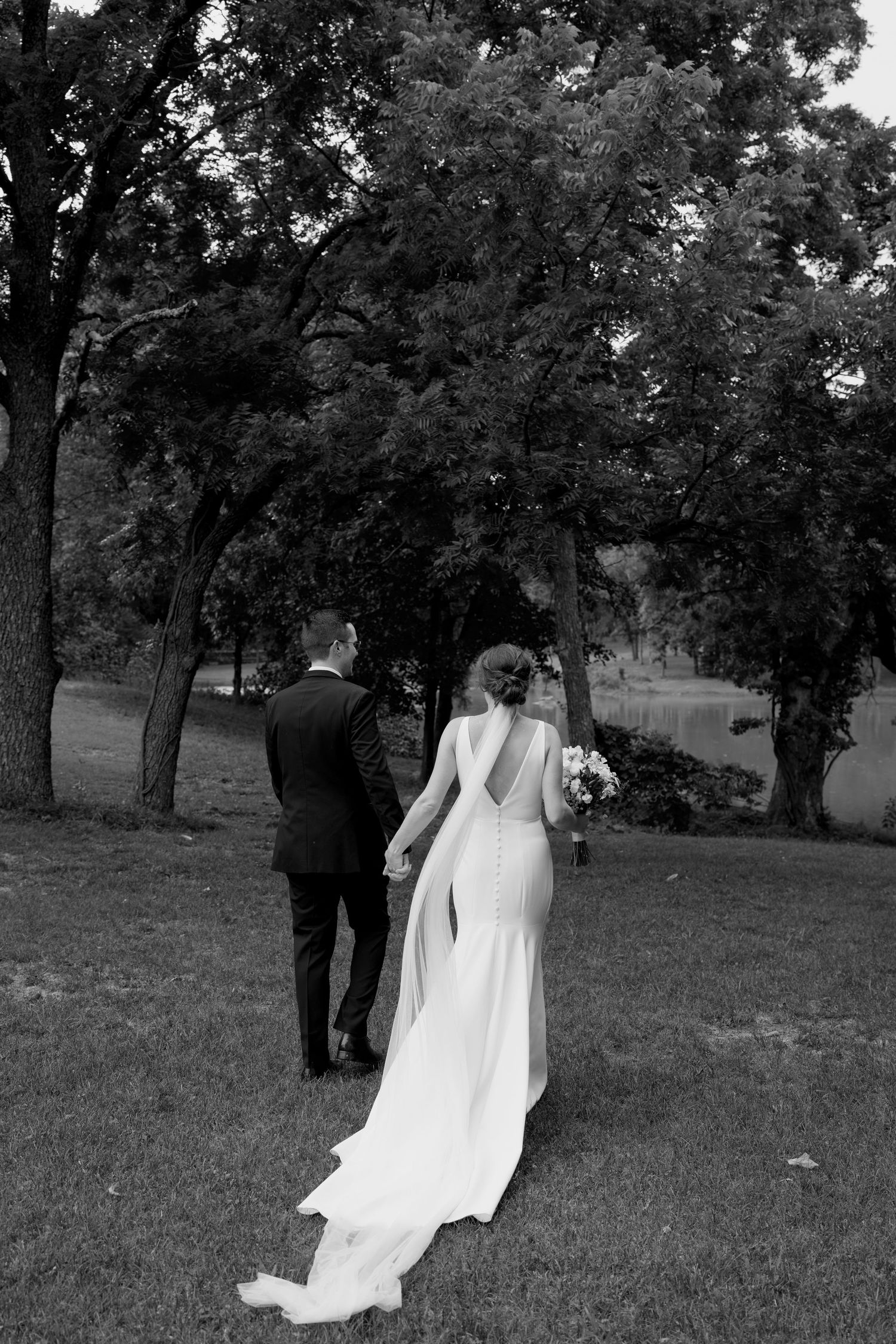 Black and white wedding photo capturing a couple walking through a tree-lined field.
