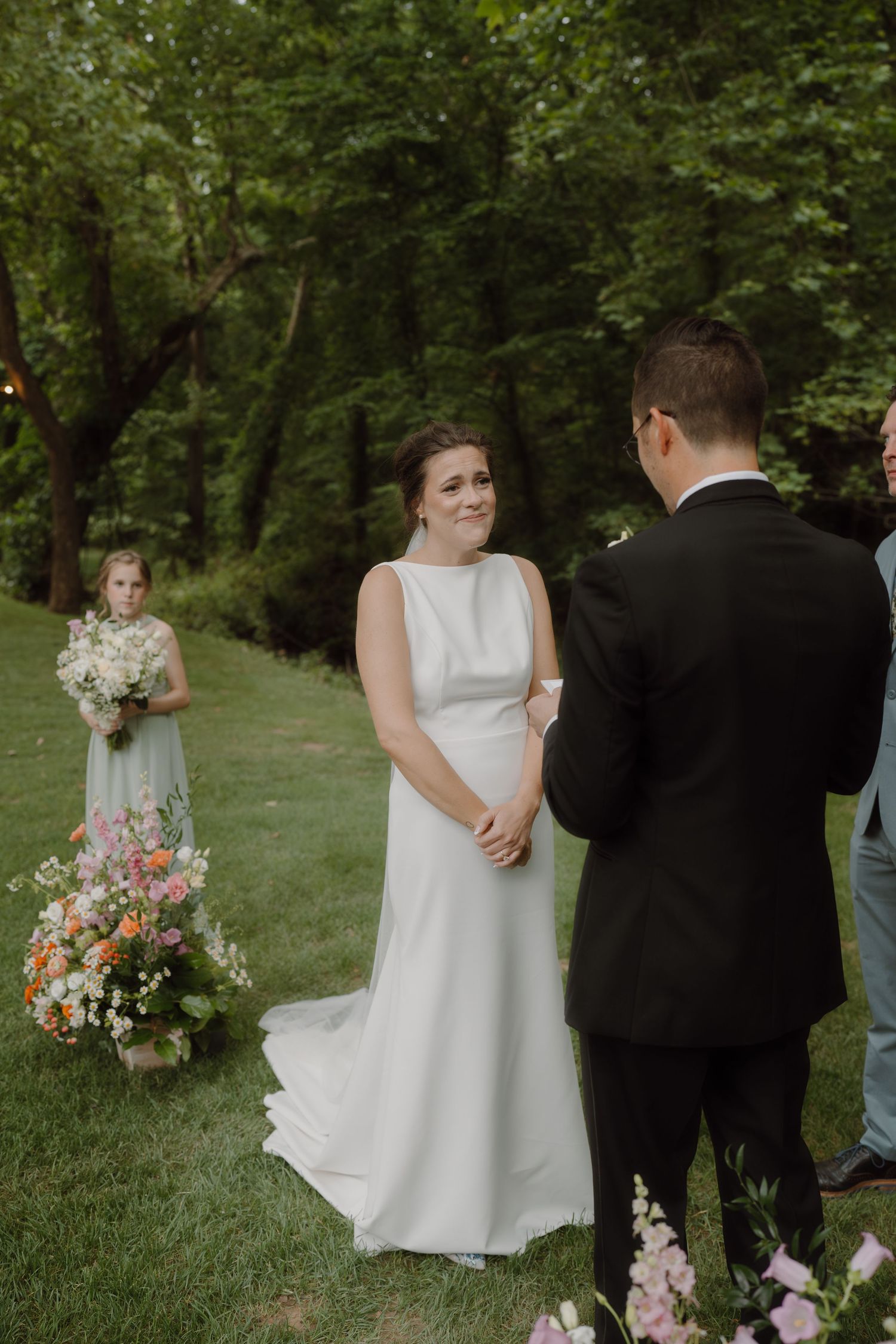 Garden wedding ceremony with white dress and colorful flower arrangements among trees.
