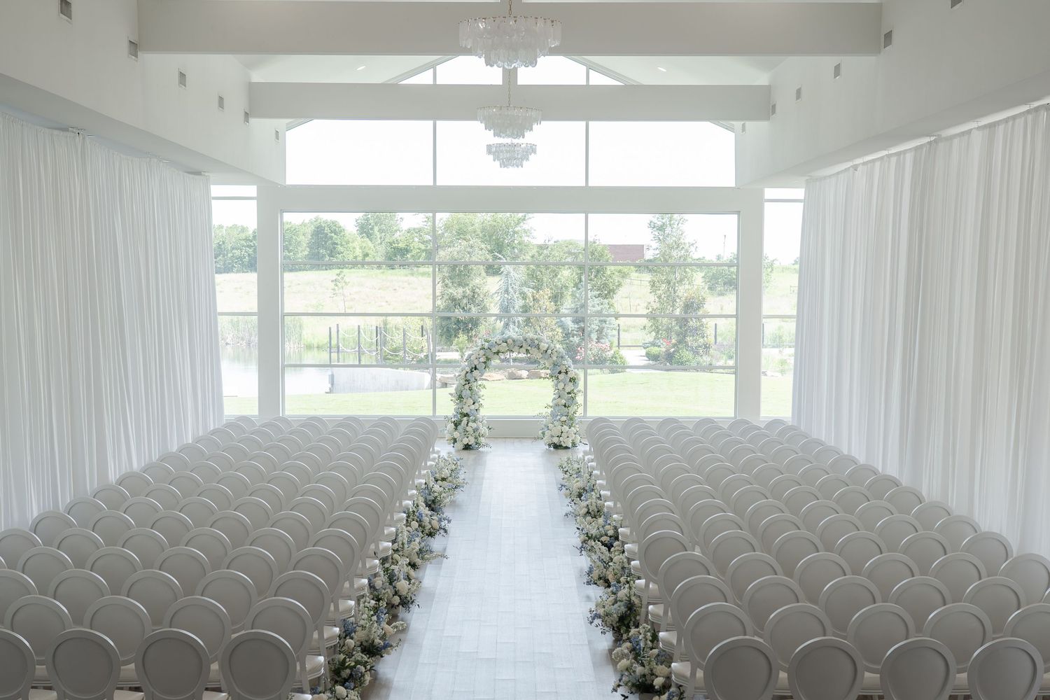 Elegant white wedding ceremony venue with rows of white chairs, floral aisle runner, and a floral arch framing garden views.