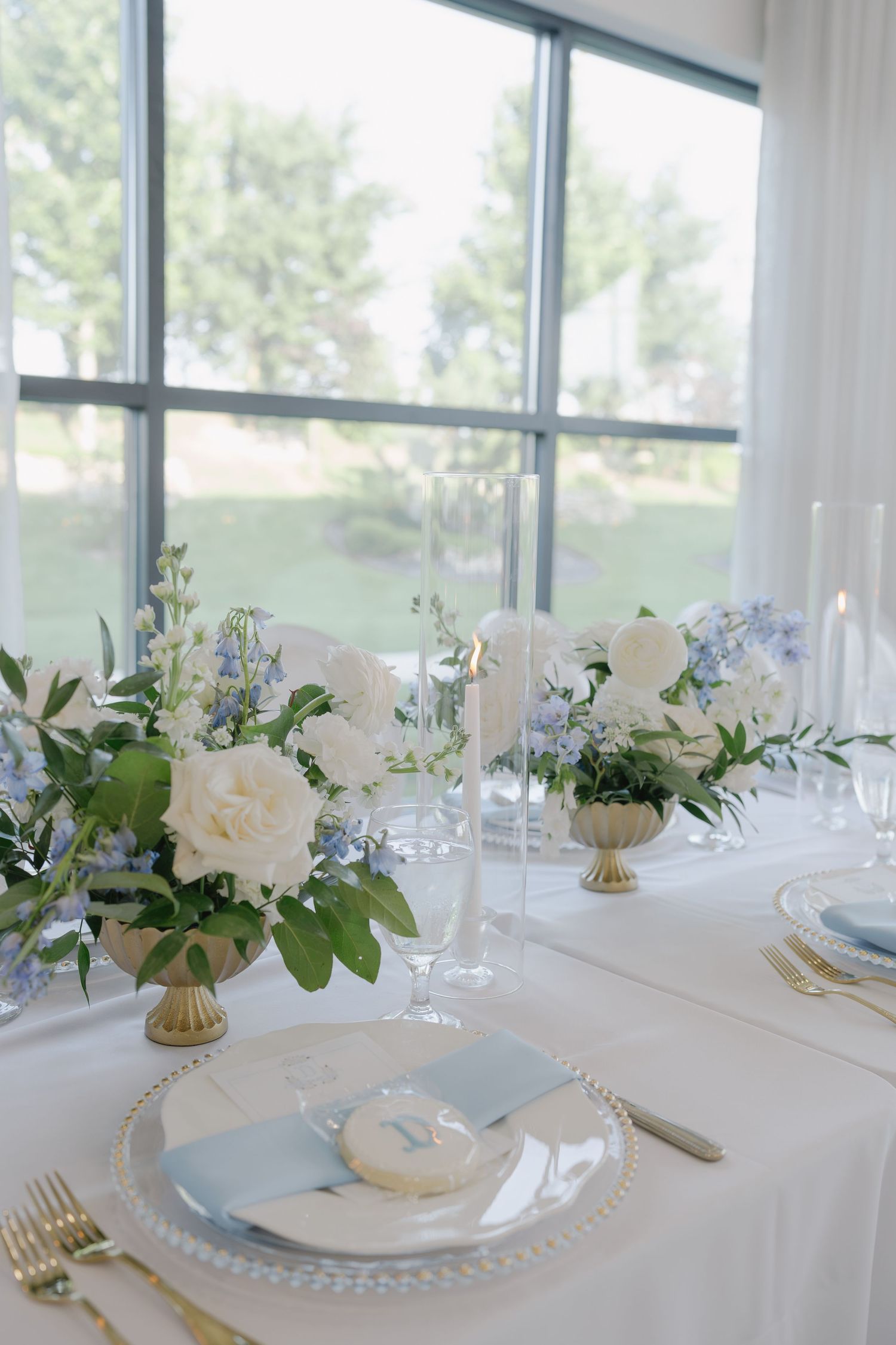 Elegant wedding table setting with white and blue floral arrangements, gold accents, crystal chargers, and tall taper candles.
