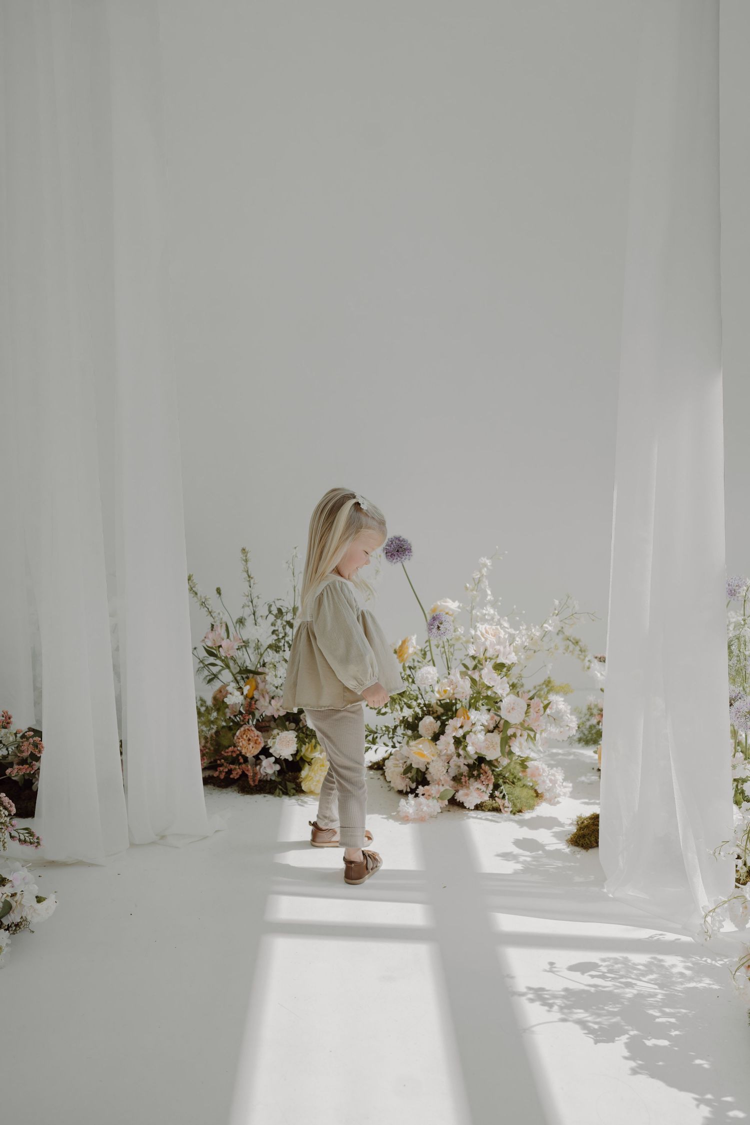 Minimalist white photography studio with scattered floral arrangements and flowing fabric creating soft shadows.