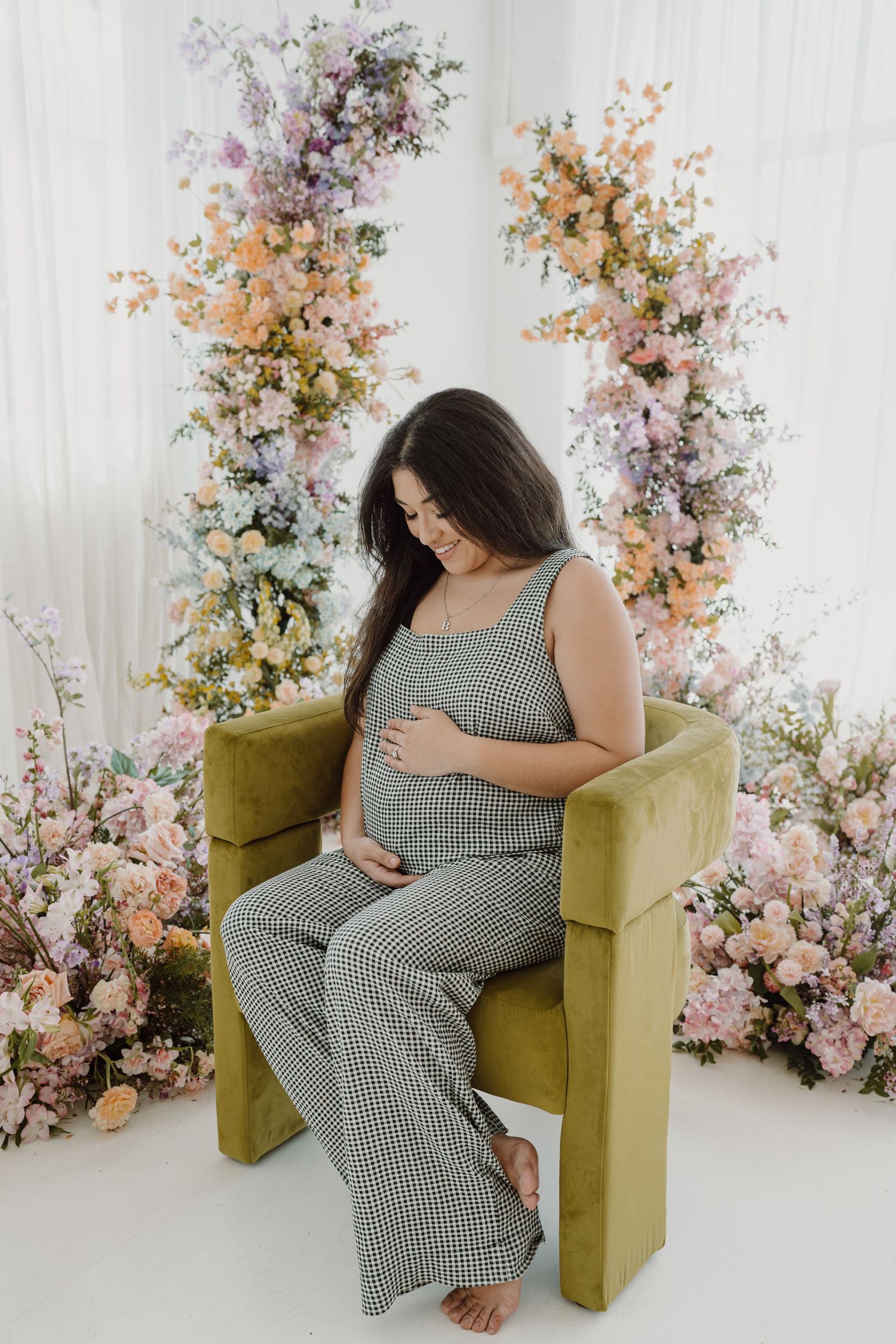 Pregnant woman in checkered jumpsuit tenderly touching belly in floral studio setting.