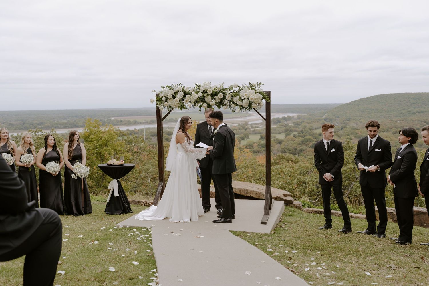Intimate wedding ceremony on hillside with wedding party and floral-adorned wooden arch.