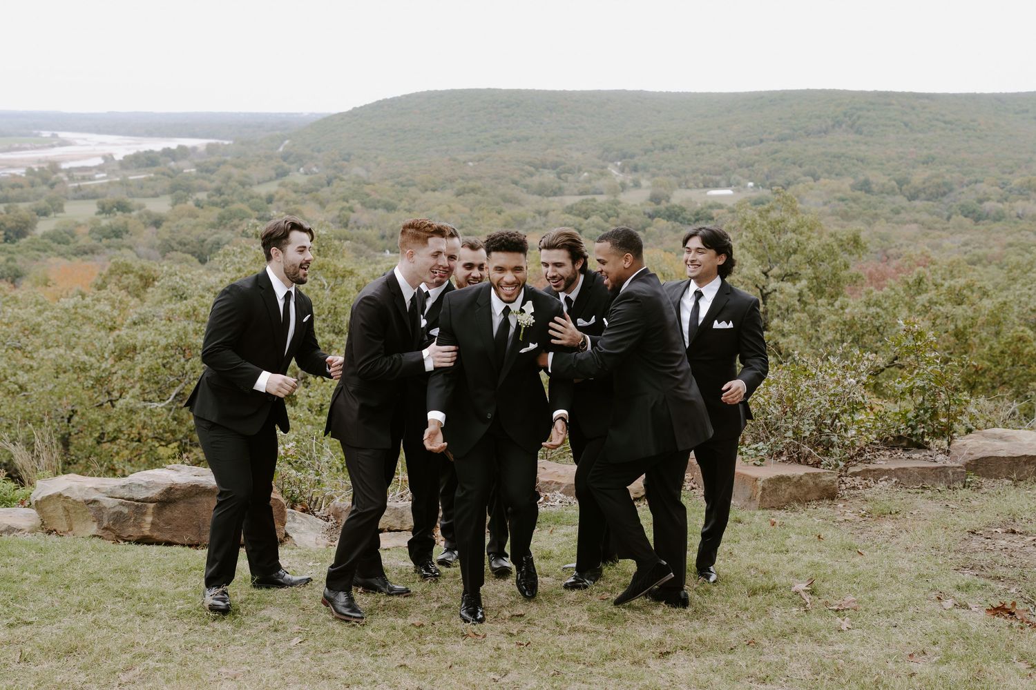 Group of groomsmen perform synchronized jumping poses during outdoor wedding photoshoot.