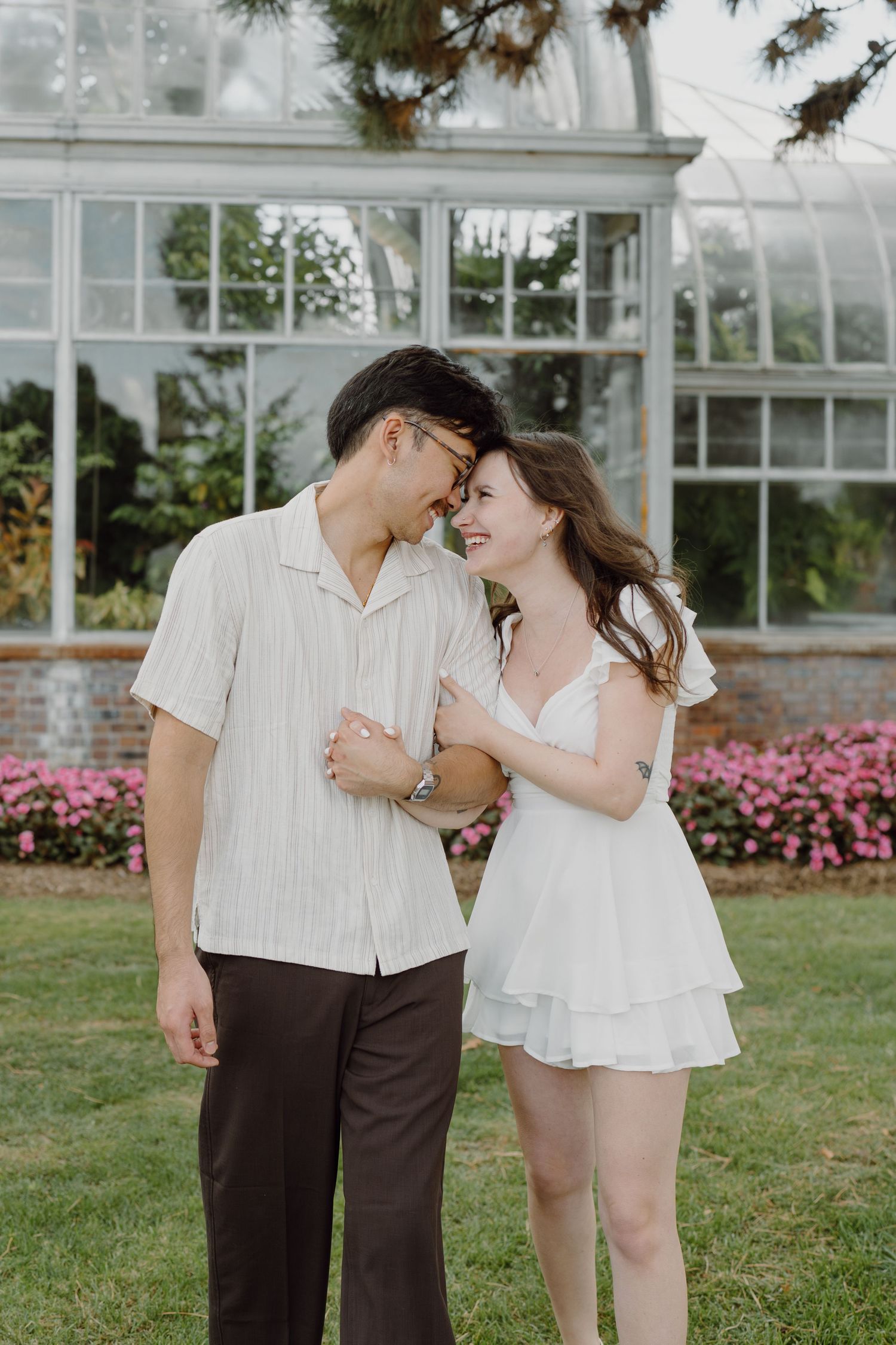 A couple embraces on a lawn with a beautiful greenhouse and pink flower garden in the background.