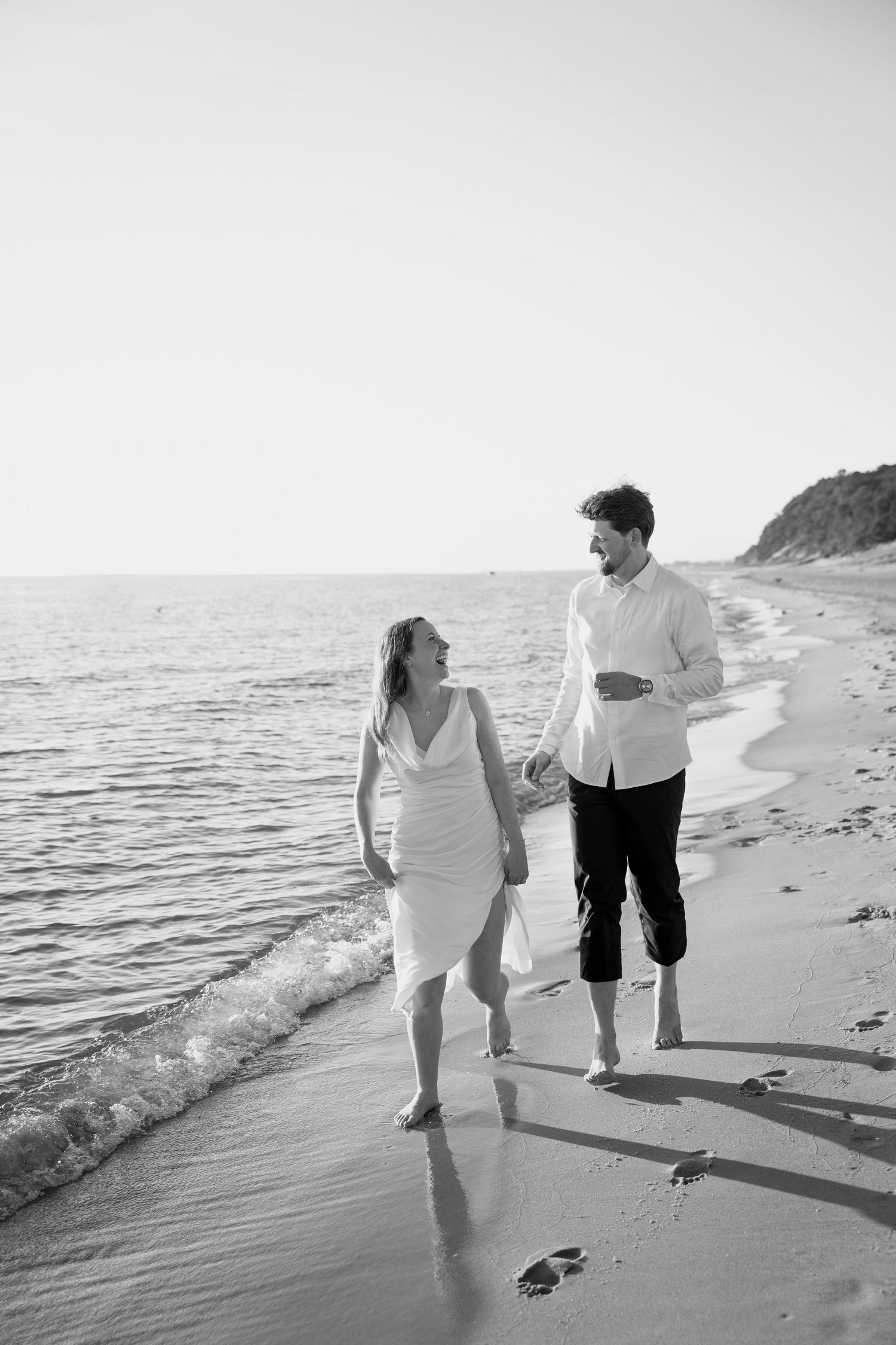 A couple walks hand in hand along the water's edge in black and white.