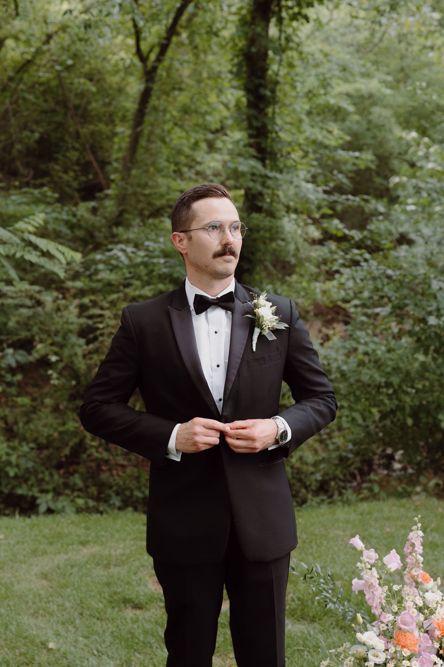A groom in a classic black tuxedo and bowtie stands in a wooded garden setting with a boutonniere.