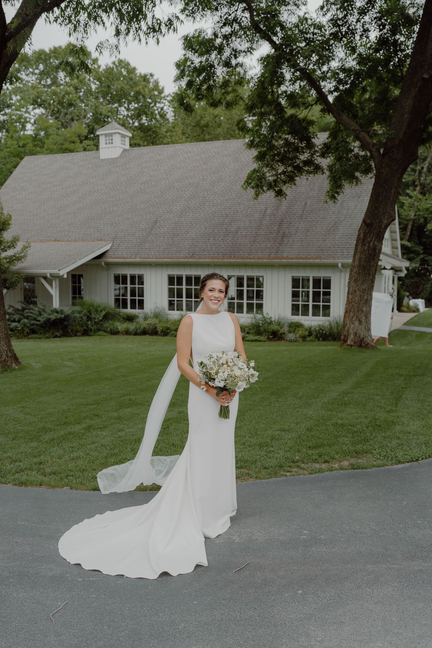 A bride in a flowing white dress walks along a curved driveway near a white building with windows.
