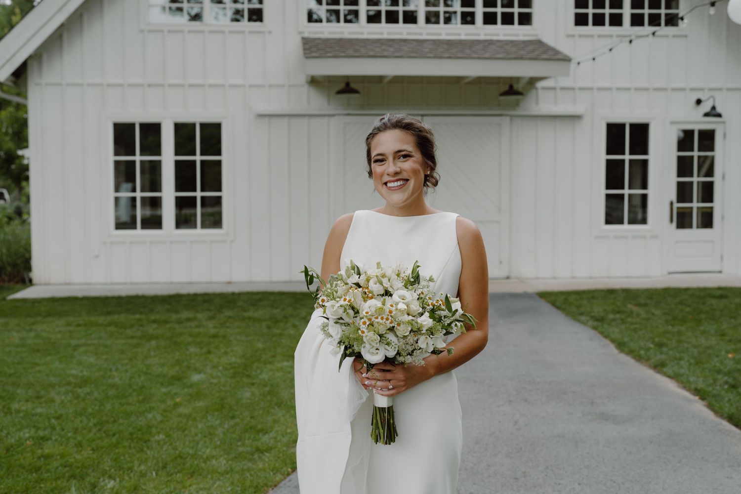 A bride poses with her bouquet against the backdrop of a white barn building with multiple windows.