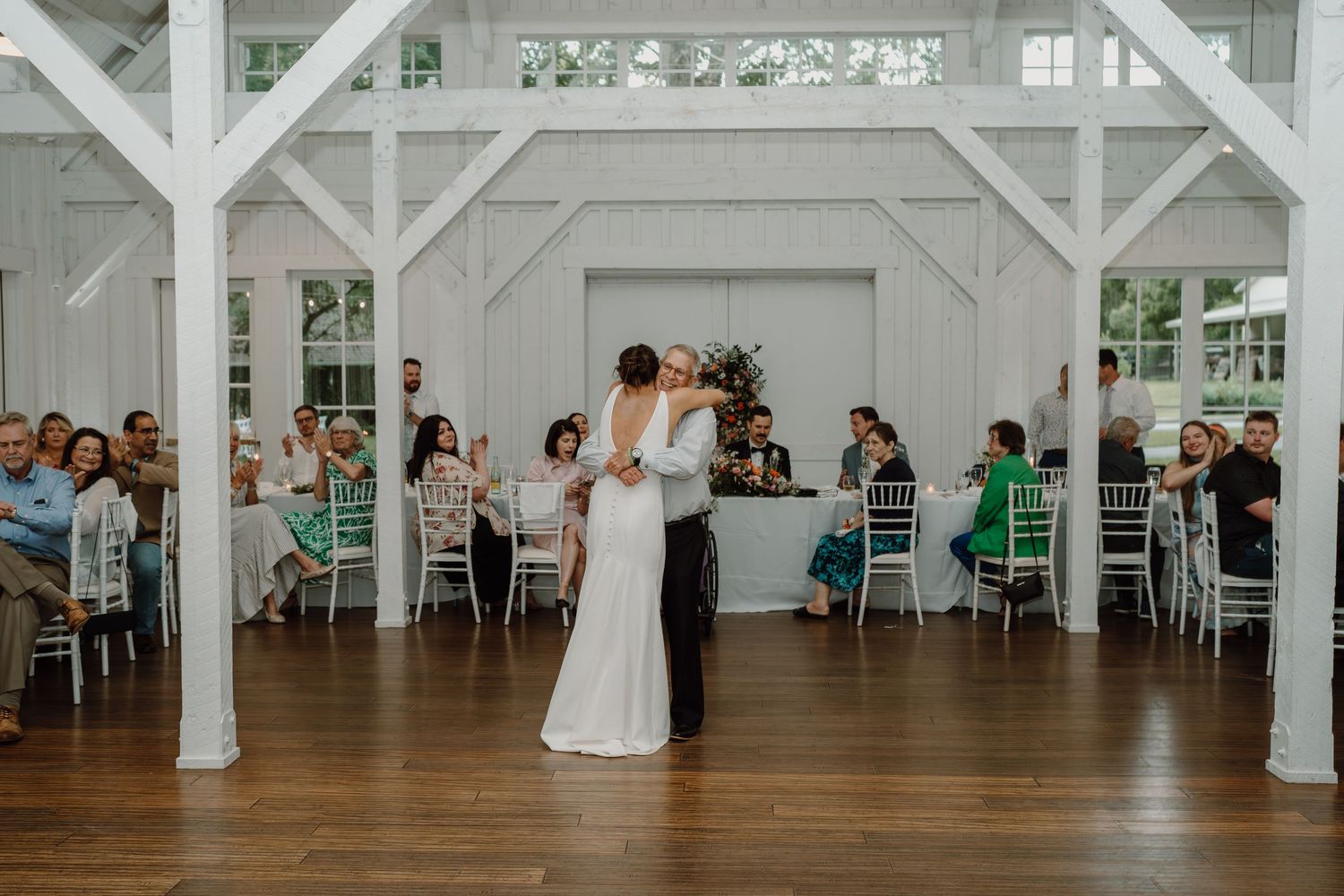 Couple dancing in the center of a bright white barn venue with guests seated at round tables.