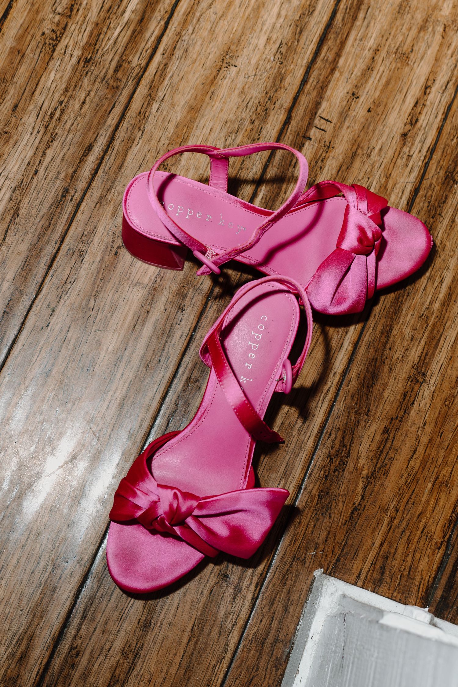 Pink satin sandals with bow details rest on a wooden floor.