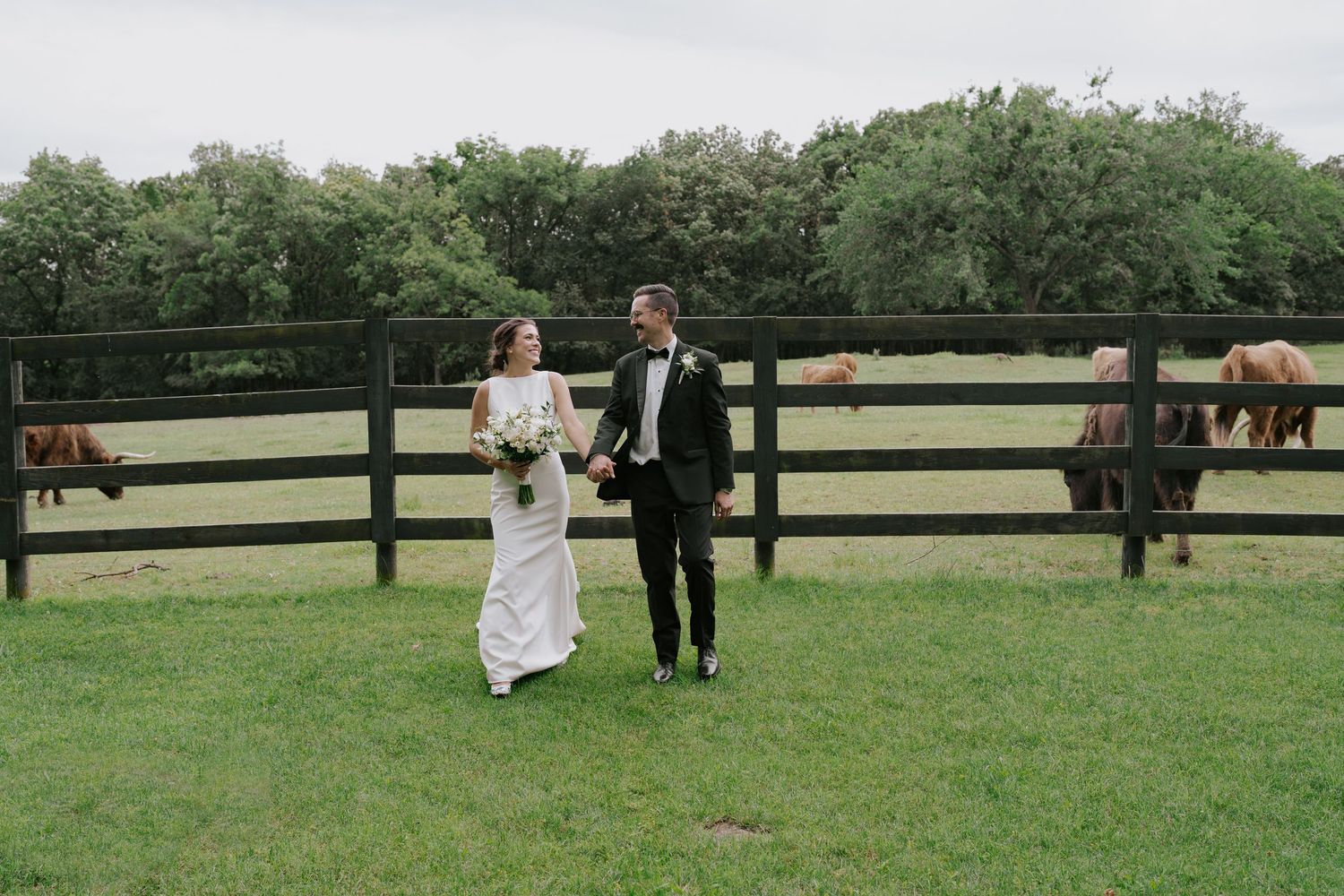 Wedding couple enjoys candid moments together on grassy field by wooden horse fence.