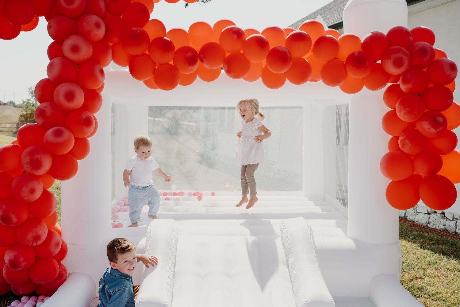 White bounce house with red balloon arch and scattered pink balls for a party.