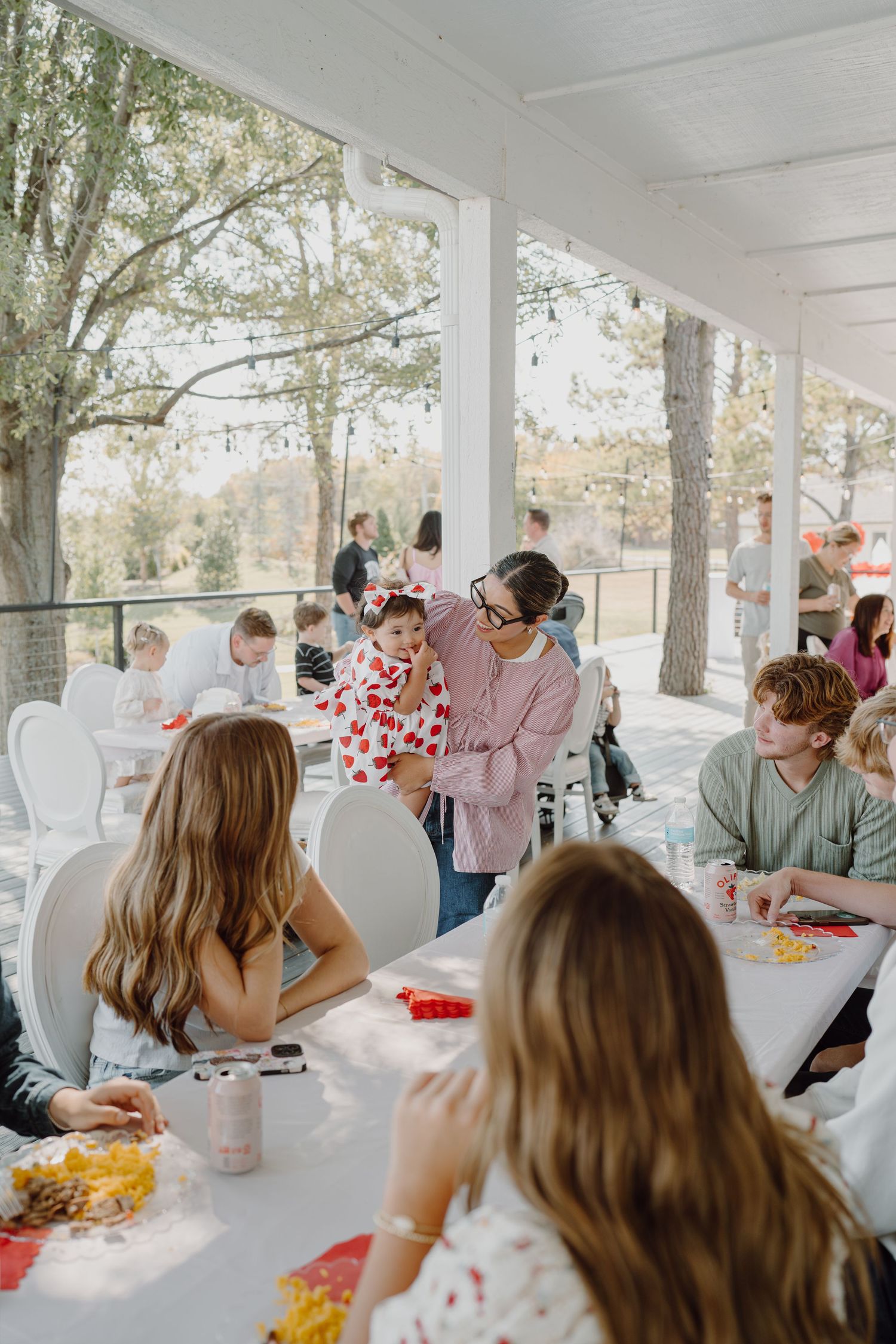 People socializing at a bright outdoor pavilion with white tables and string lights overhead.