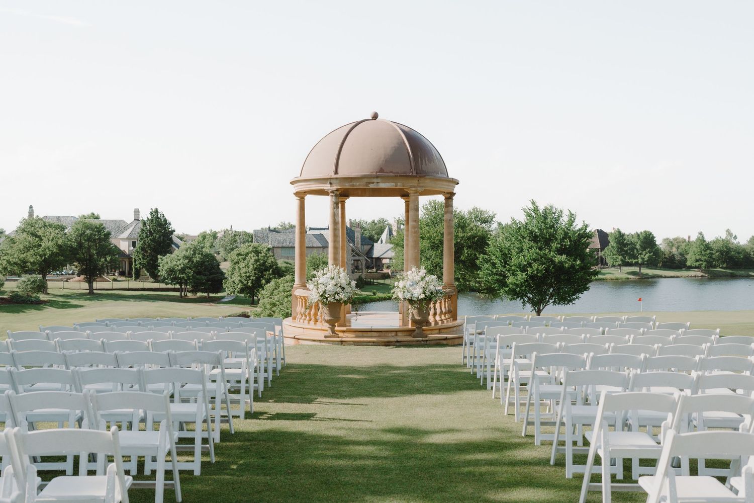 Elegant outdoor wedding ceremony setup with white chairs facing a domed gazebo overlooking a serene lake and green landscape.
