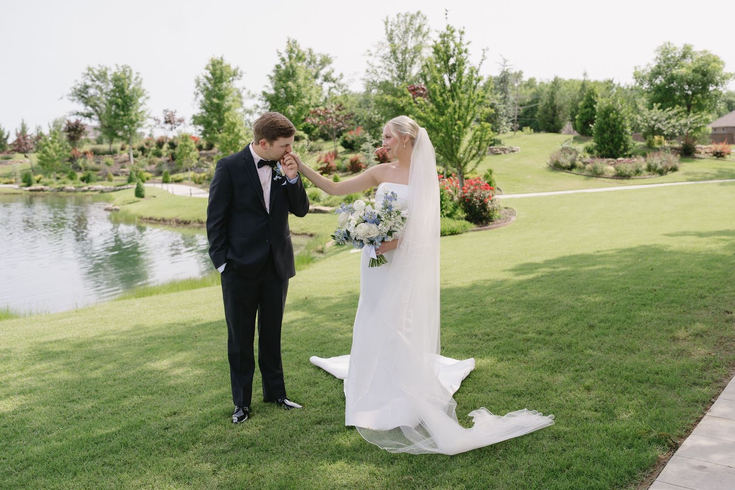 Groom in black tuxedo kisses bride's hand outdoors near pond, bride holds blue and white bouquet in elegant white gown.