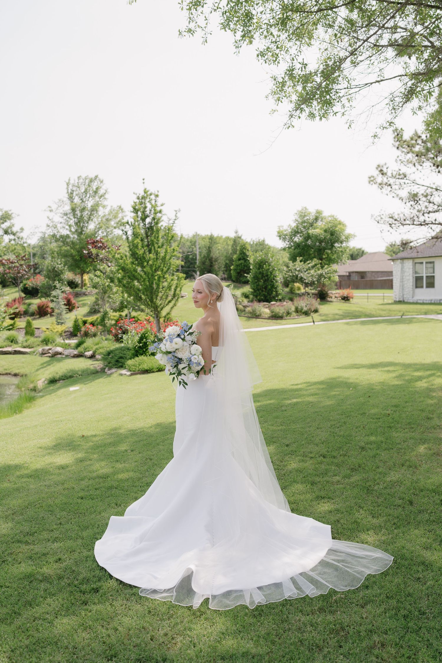 Bride in white gown with long veil holds bouquet on lush green lawn surrounded by garden landscaping.