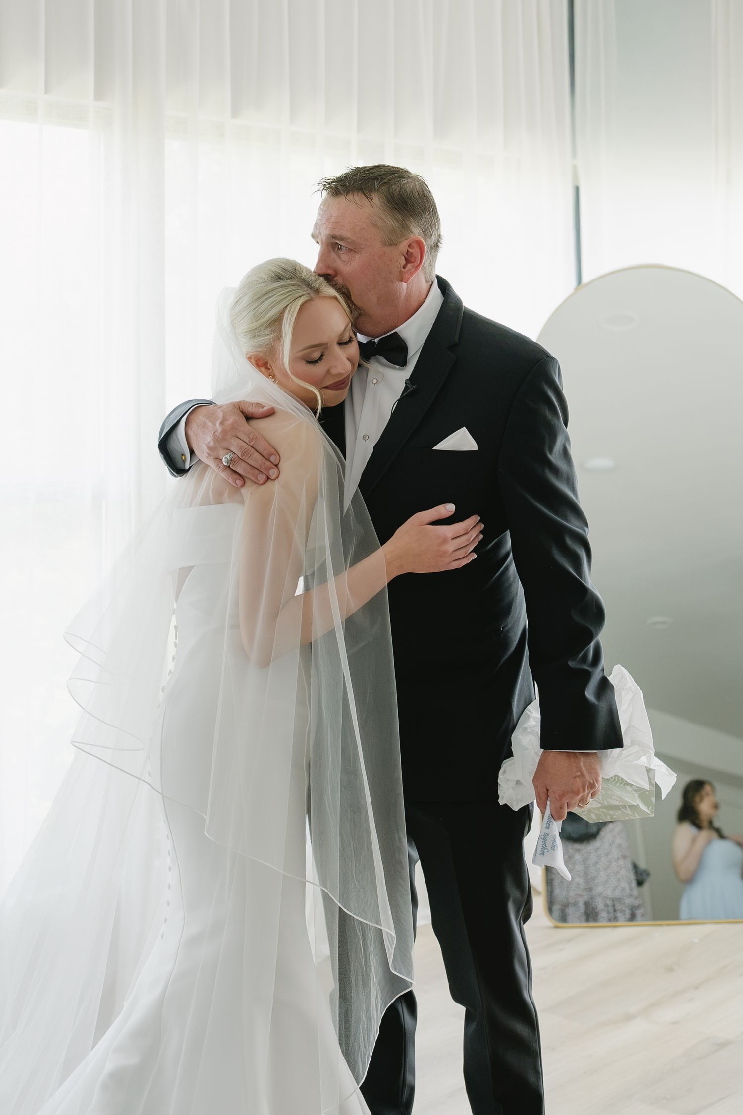 A bride in a white gown and veil embraces a man in a black tuxedo sharing a tender moment indoors.