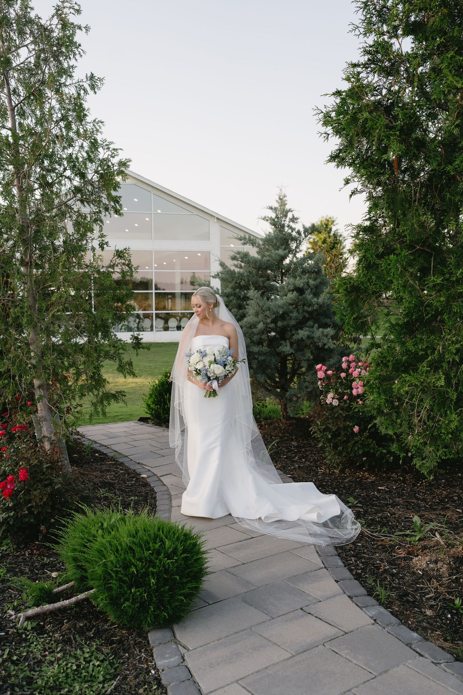 Bride in white gown and veil holds bouquet on stone path surrounded by lush garden greenery near elegant venue.