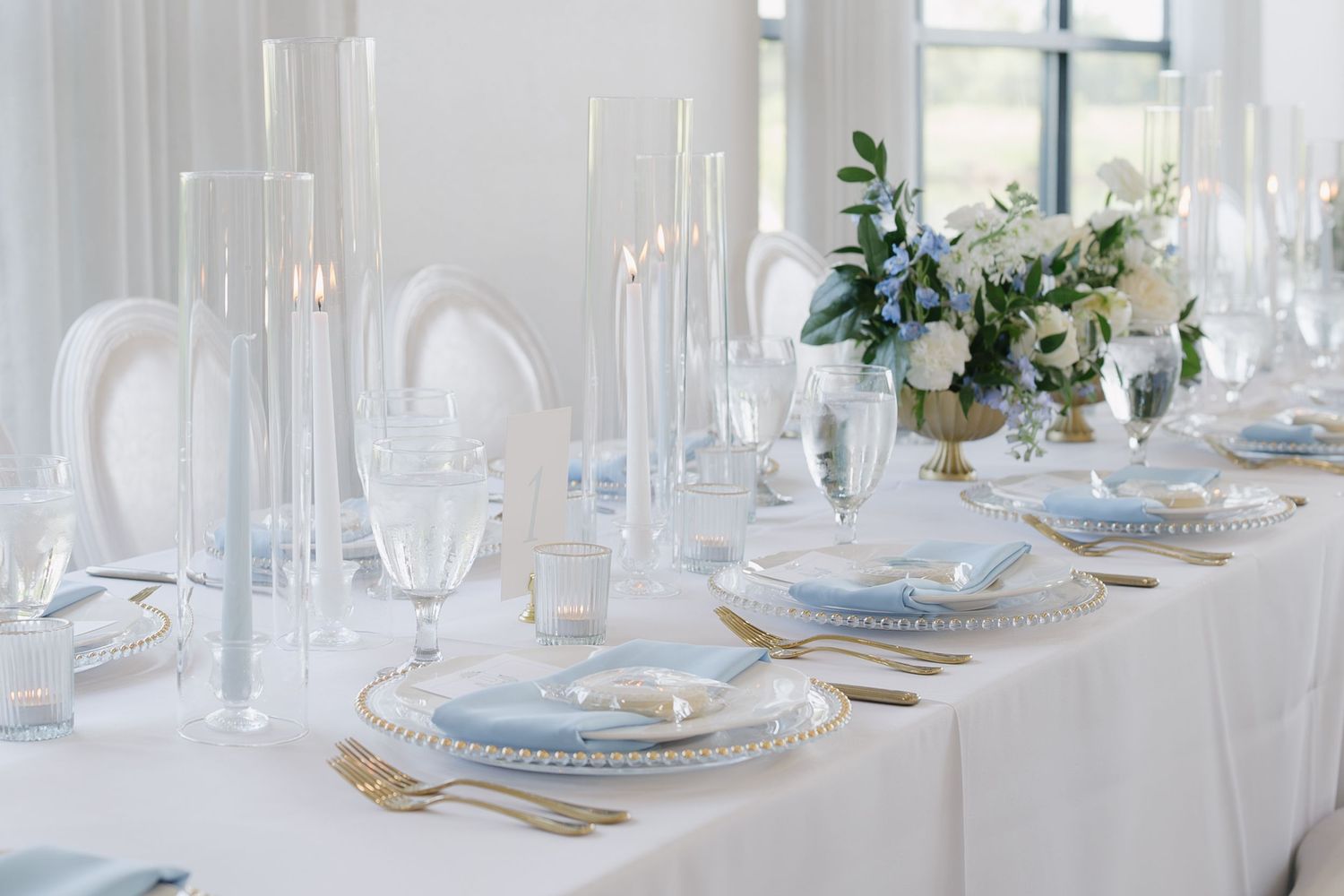 Elegant wedding table setting with white linens, gold cutlery, blue-rimmed plates, tall candles, and white floral centerpiece.