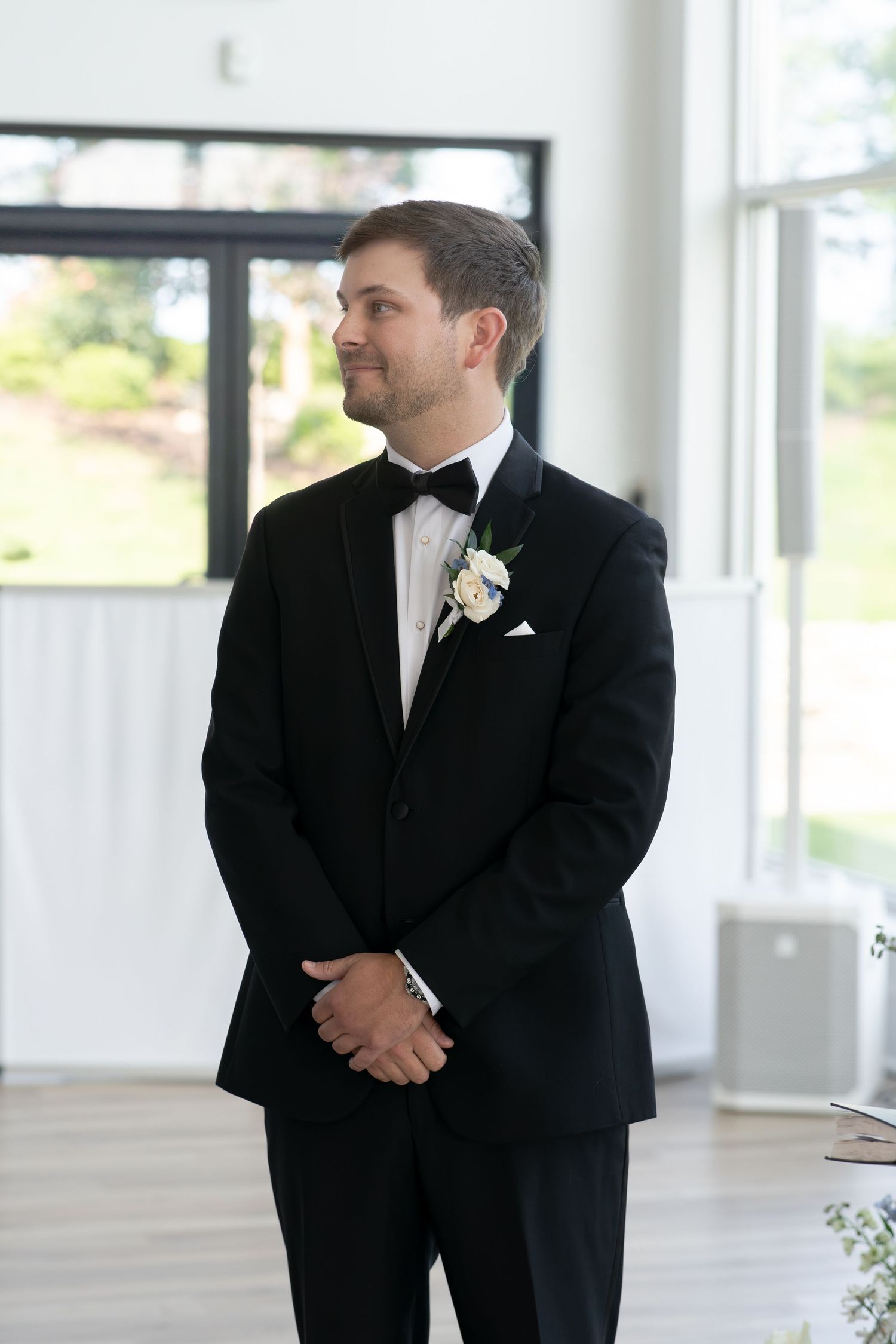 Groom in black tuxedo with white boutonniere and bow tie standing in bright wedding venue.