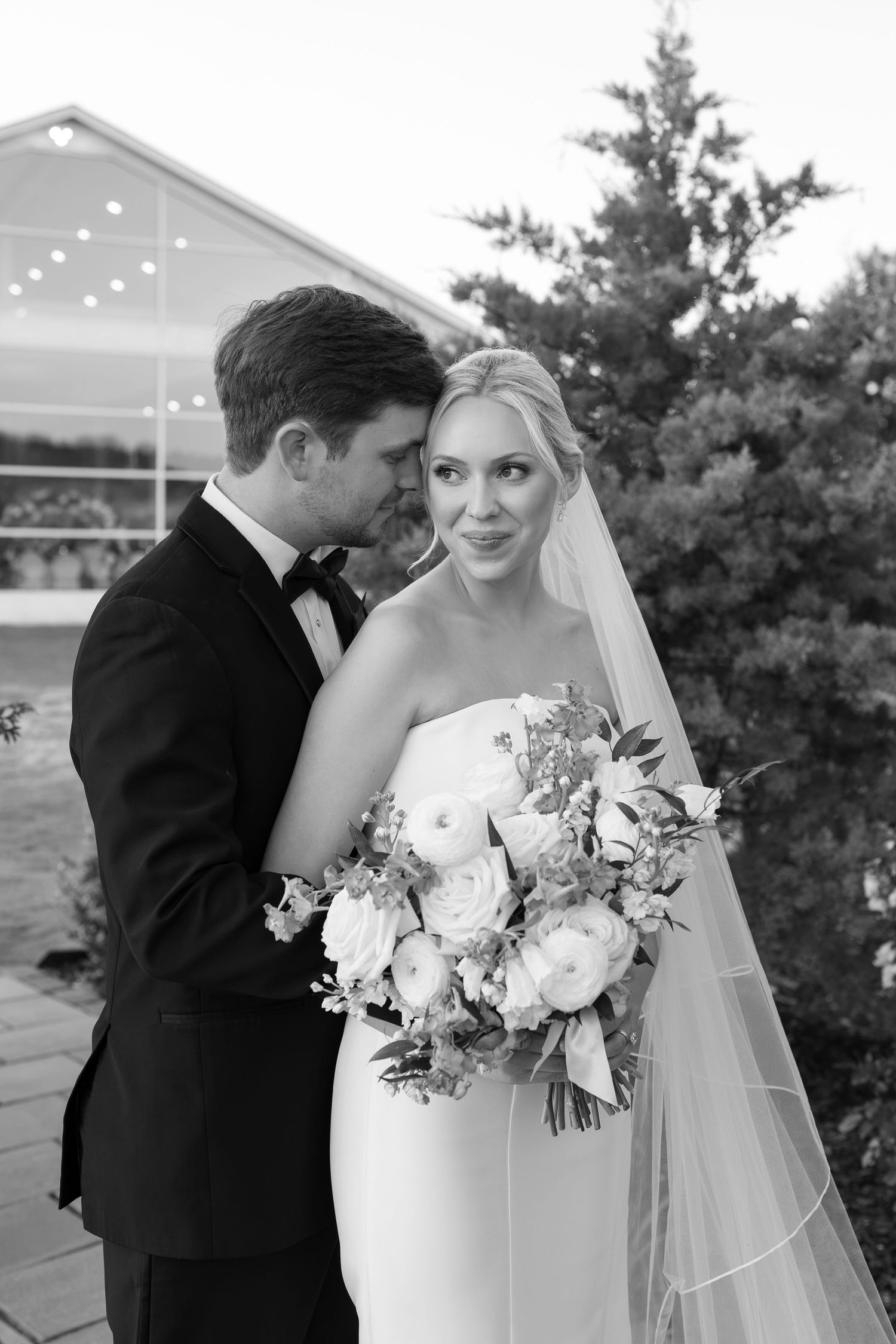 Black and white wedding portrait of bride holding bouquet and groom in tuxedo embracing outdoors near greenhouse.