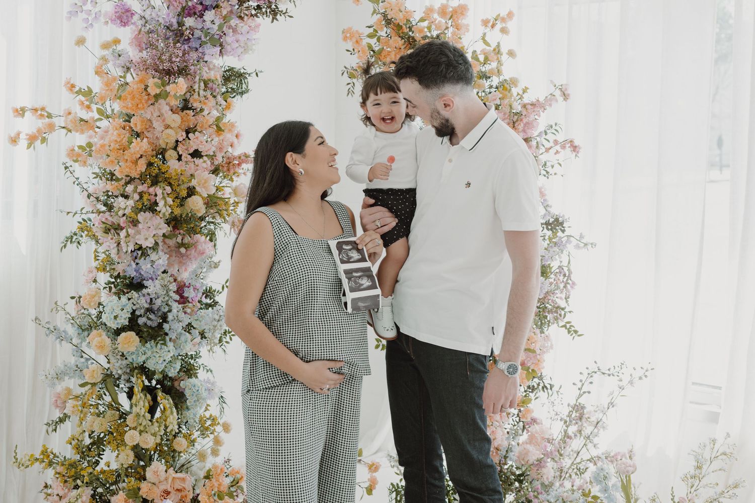 A couple shares a joyful moment surrounded by colorful floral arrangements in a bright studio setting.