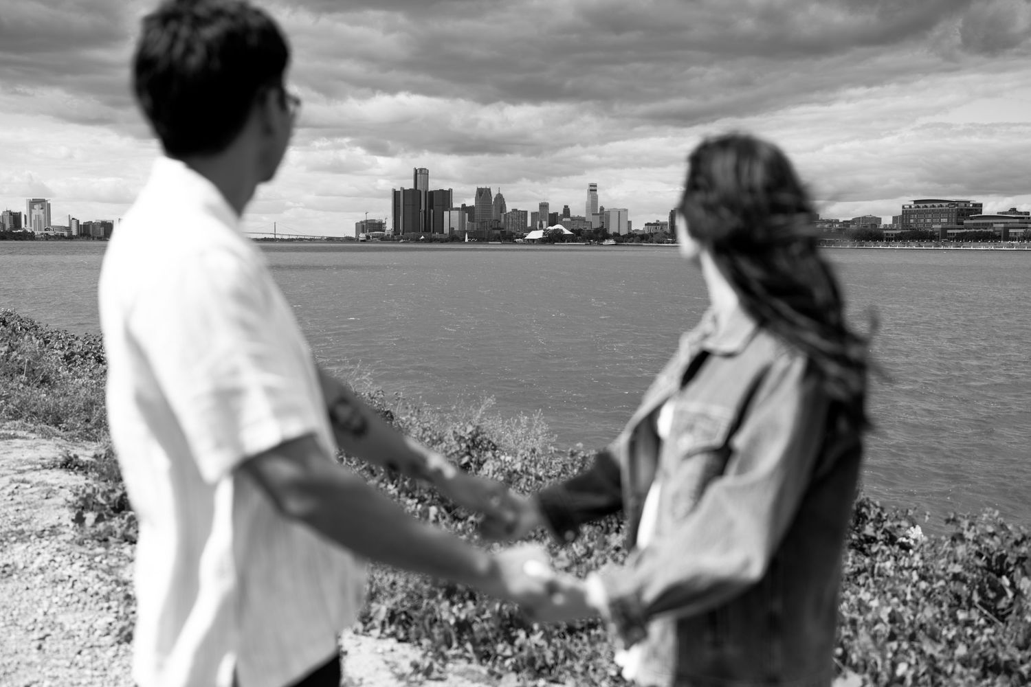 Black and white photo of couple holding hands while overlooking city skyline across water.