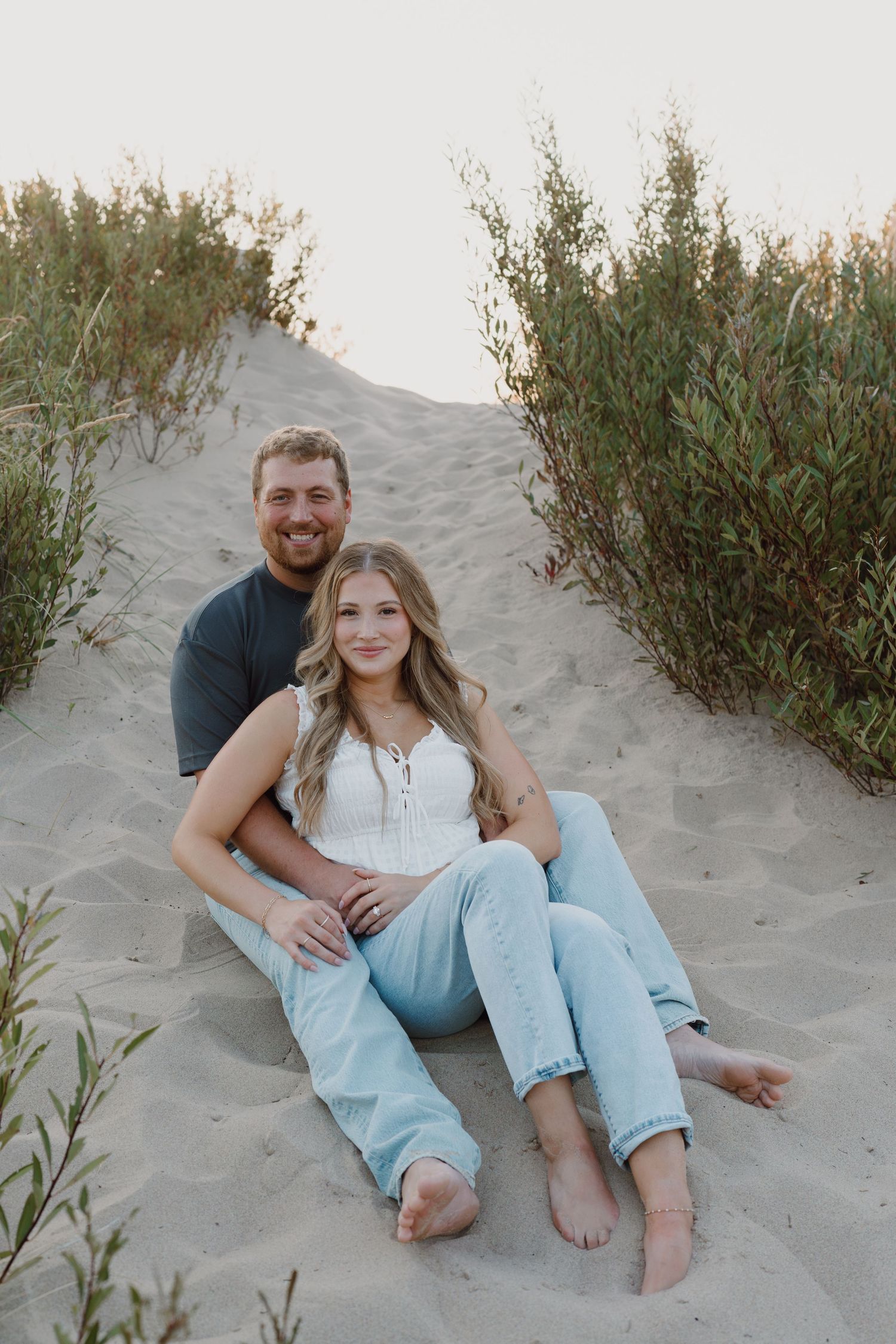 A couple relaxes on sandy beach dunes surrounded by coastal vegetation.